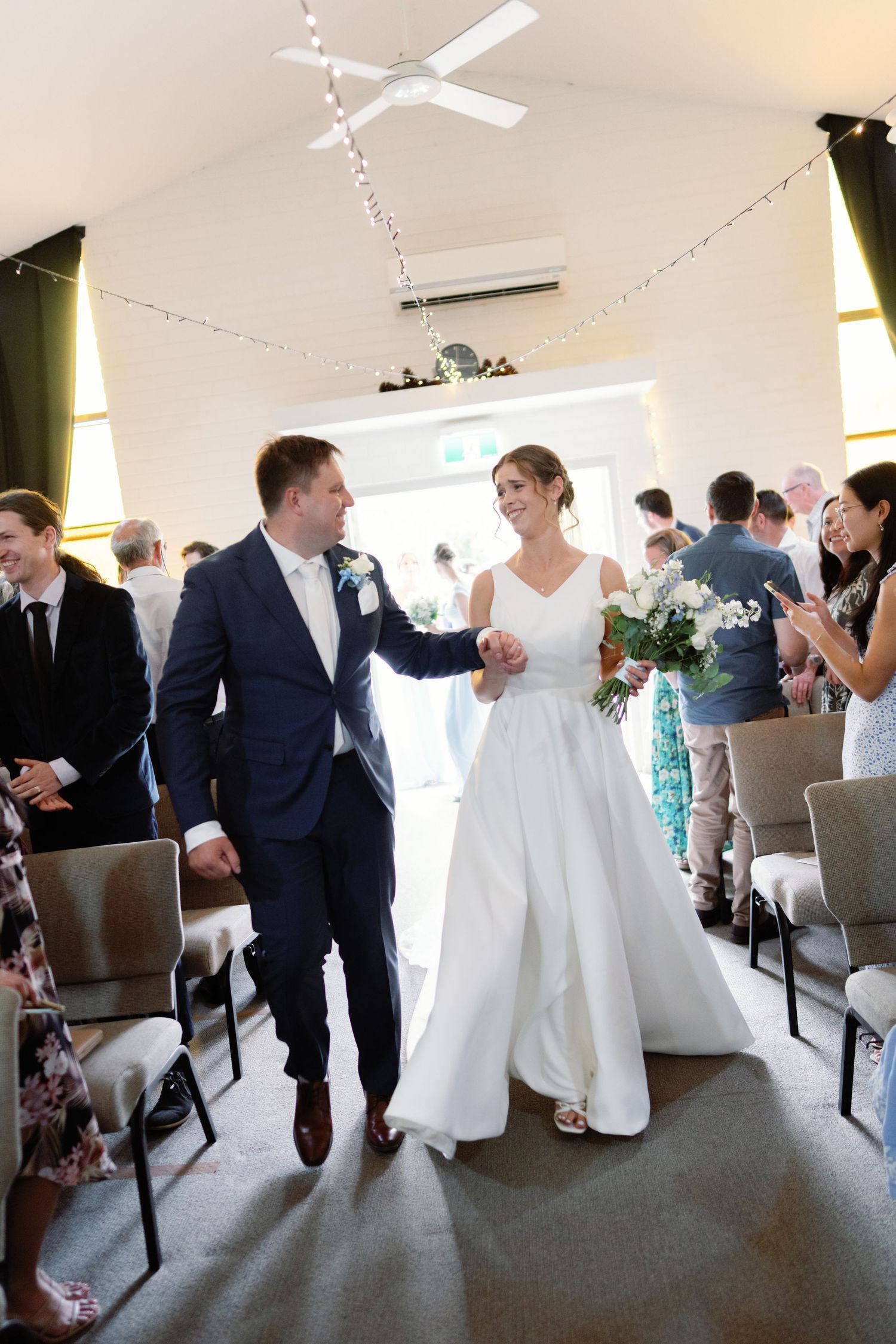 Wedding reception scene with guests mingling in a bright indoor venue decorated with string lights.