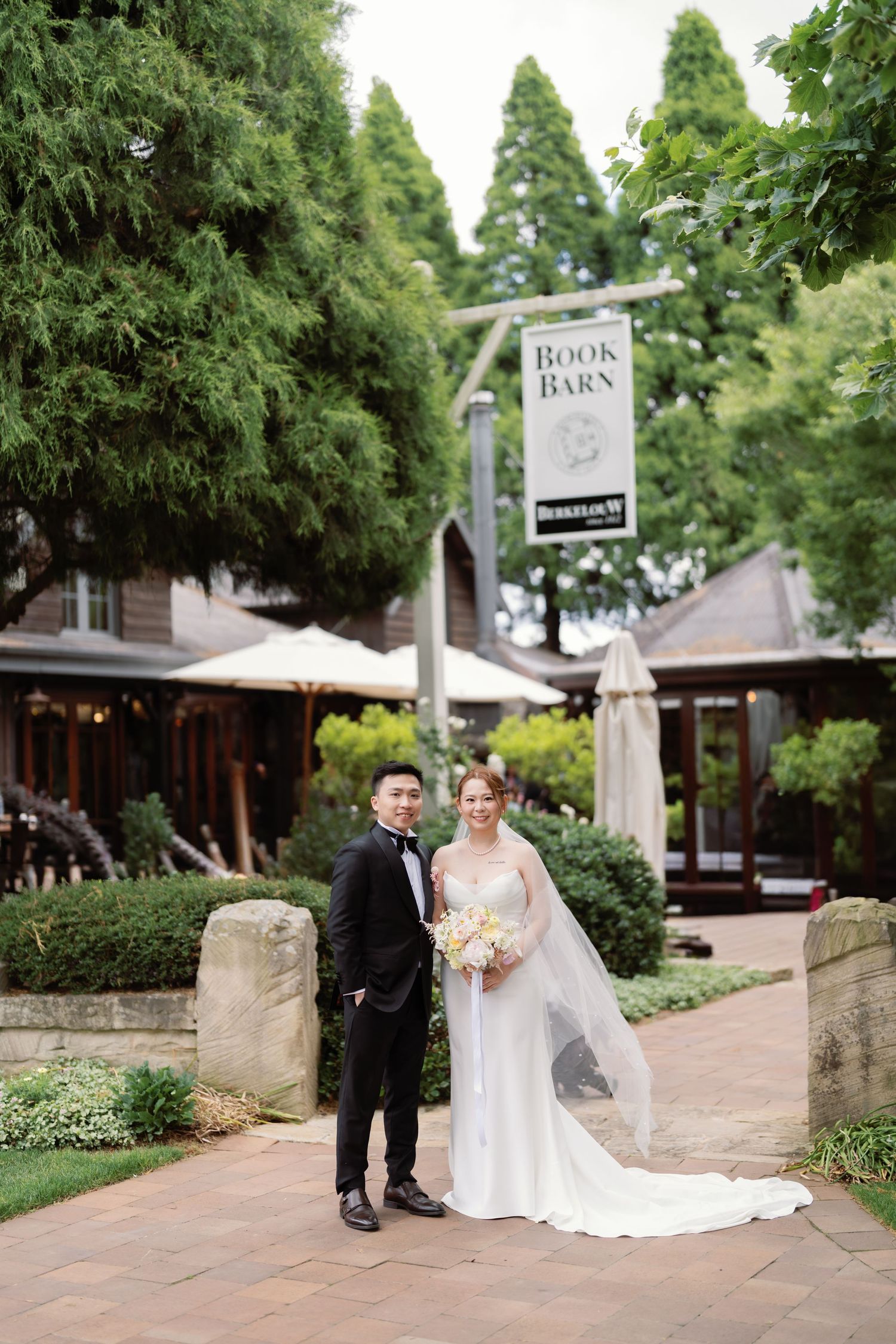 Wedding couple poses along a stone pathway surrounded by lush green trees and gardens.