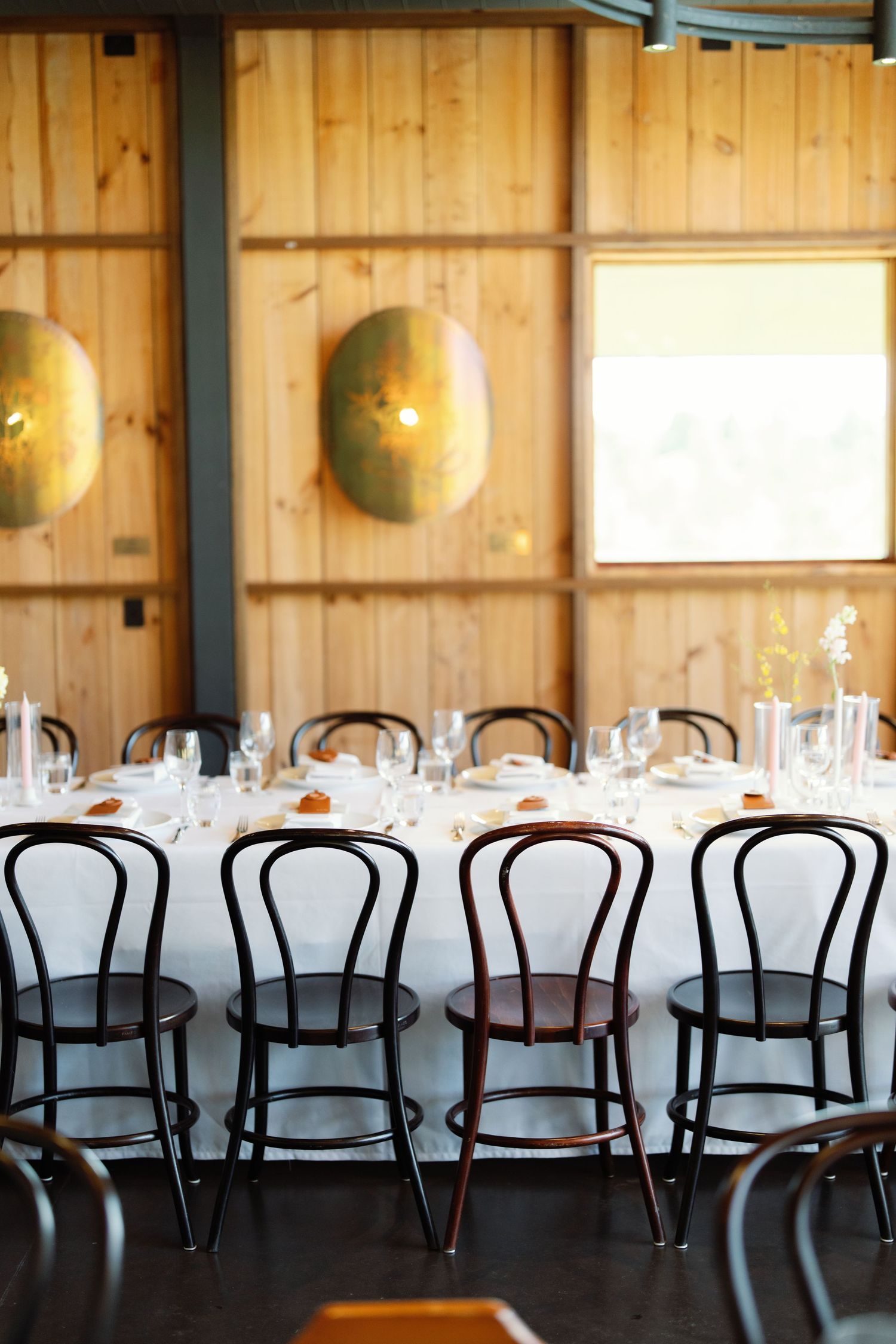 Long banquet tables with white linens and dark bentwood chairs set up in a rustic wooden room with wall sconces.