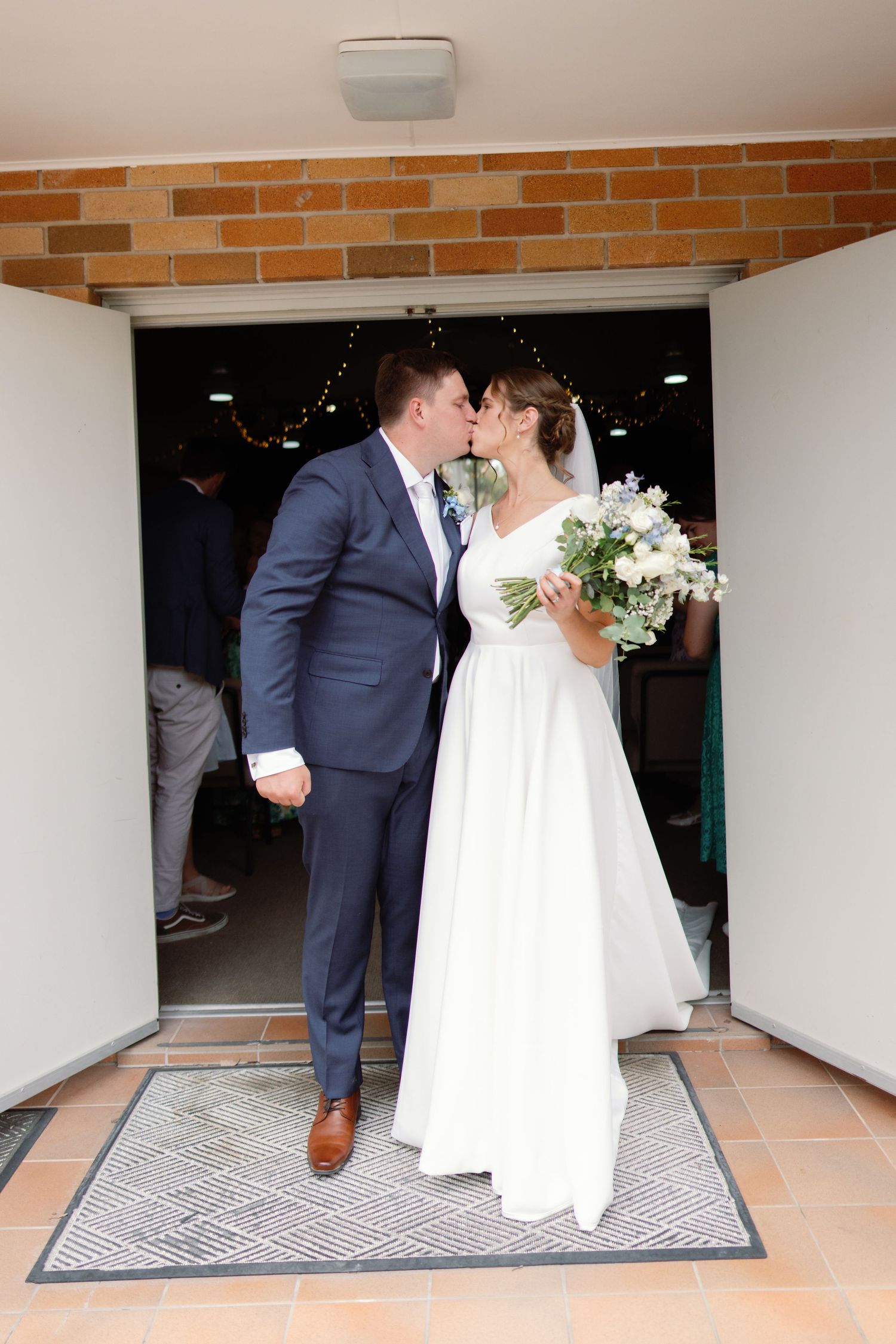 Wedding couple in formal attire walking through covered entrance with brick walls.