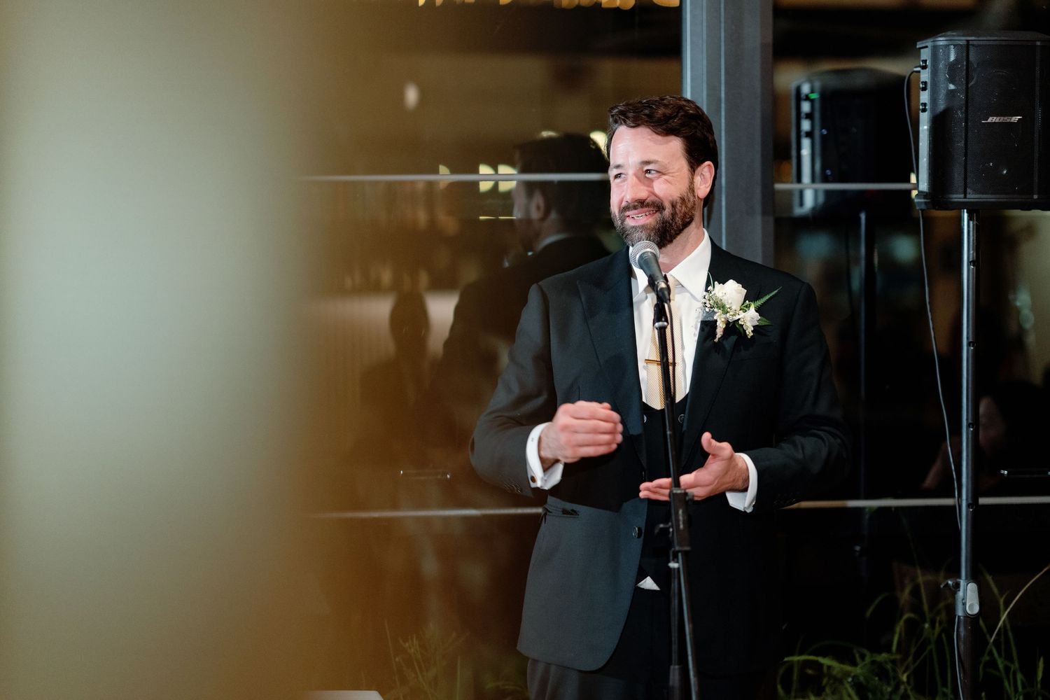 Smiling groom in black suit with boutonniere delivers speech at microphone during wedding reception.