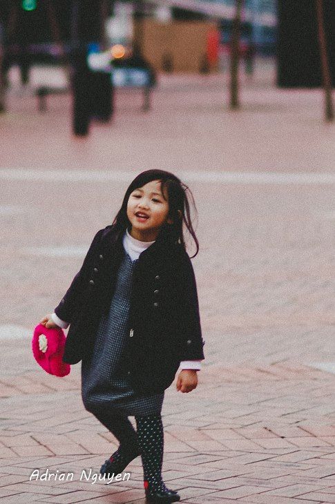 Child in a black coat walking on brick pavement in an urban setting.