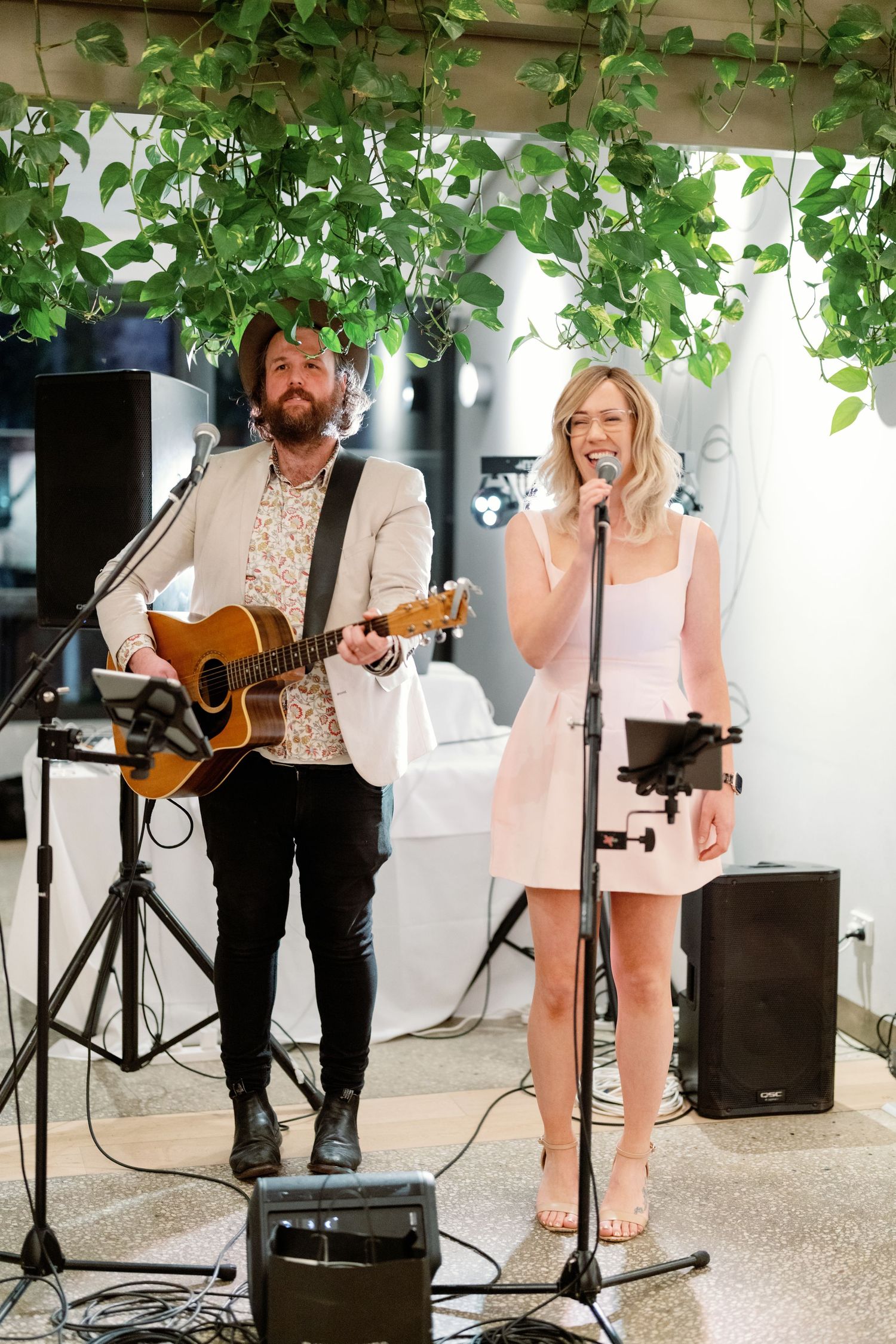 Live wedding band duo performing with acoustic guitar and vocals under hanging greenery at indoor reception venue.