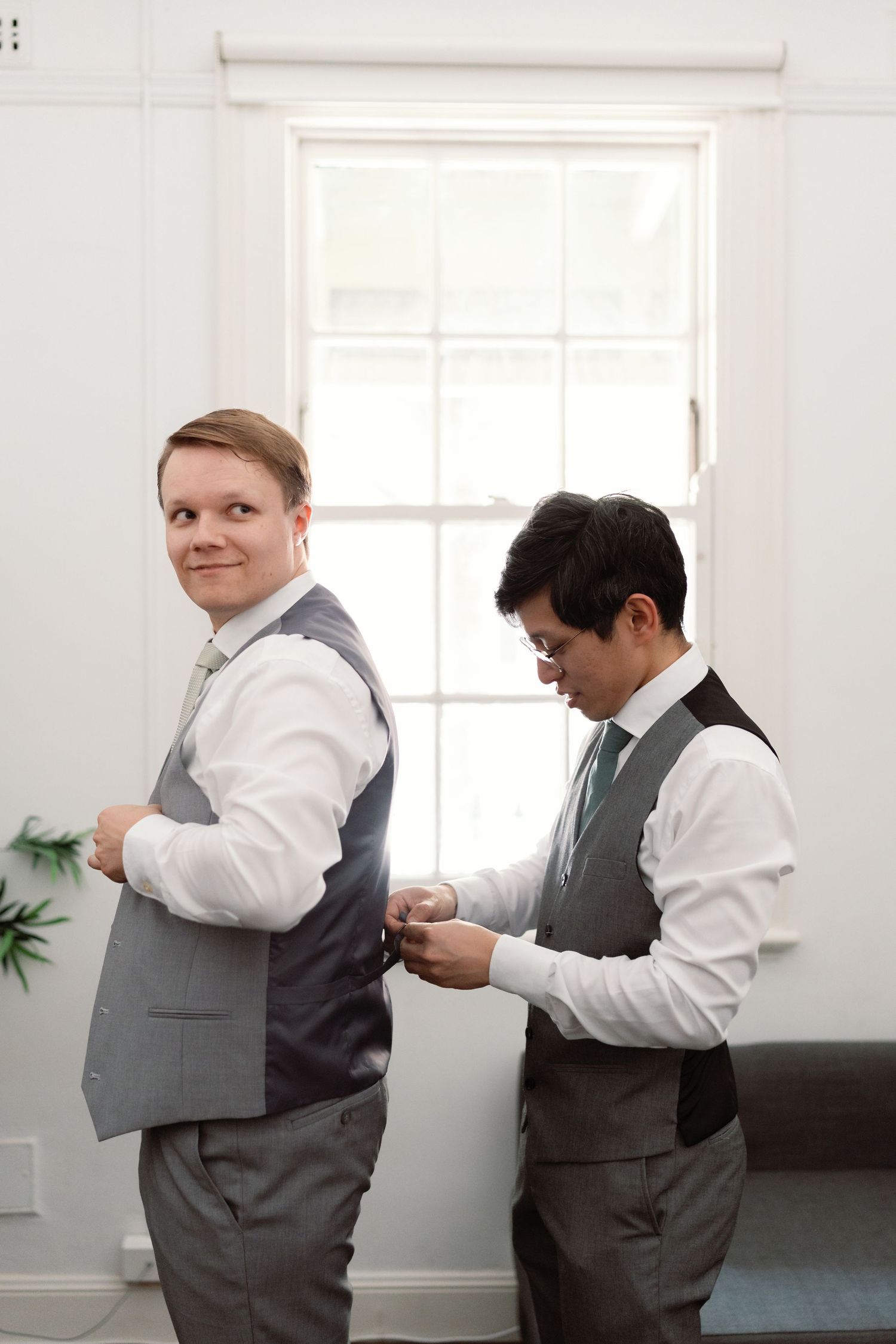 Two groomsmen in matching gray vests and white shirts getting ready before a wedding ceremony by a bright window.