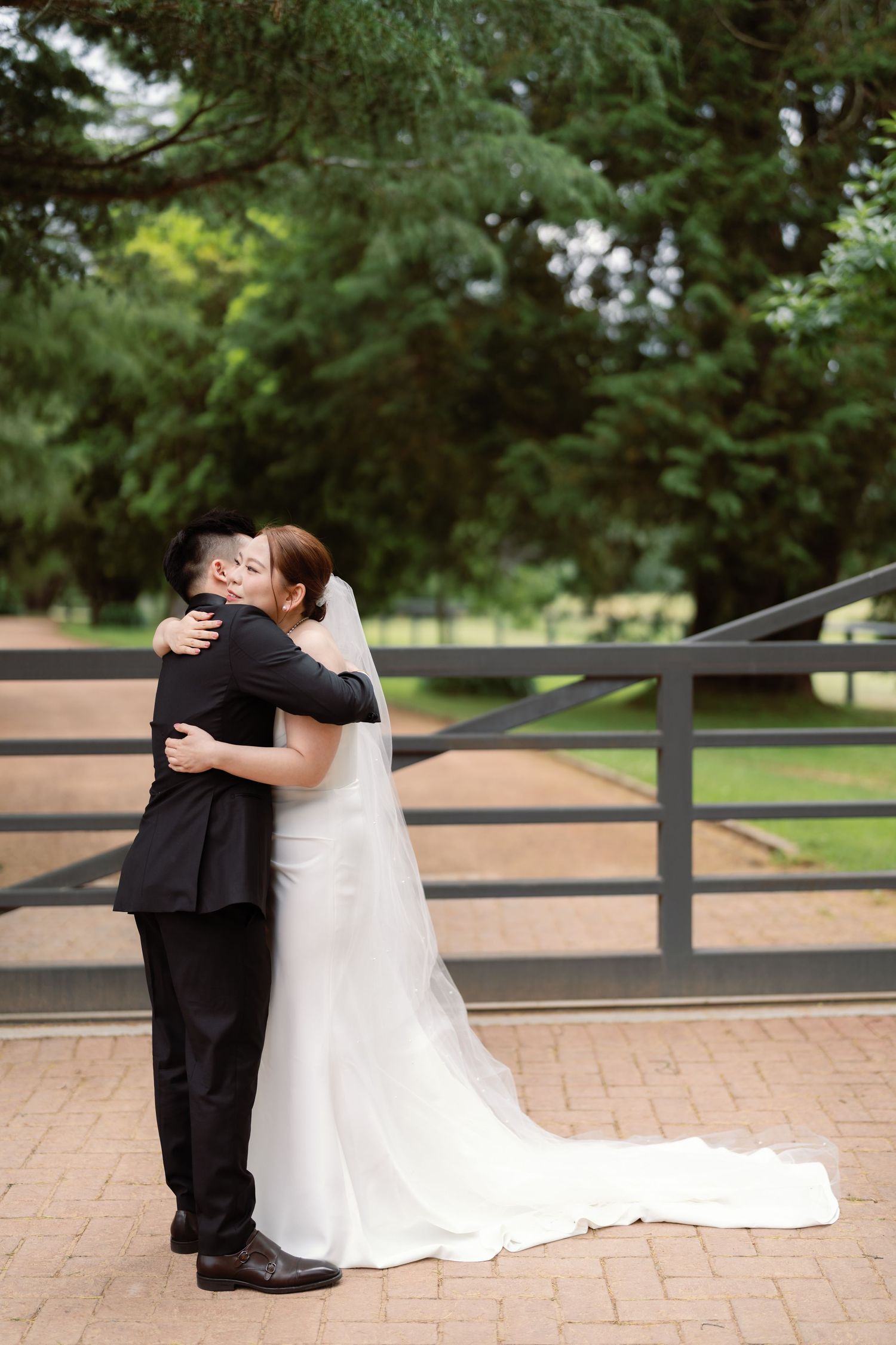A romantic first look moment between newlyweds on a brick patio with black fencing and evergreen trees in background.