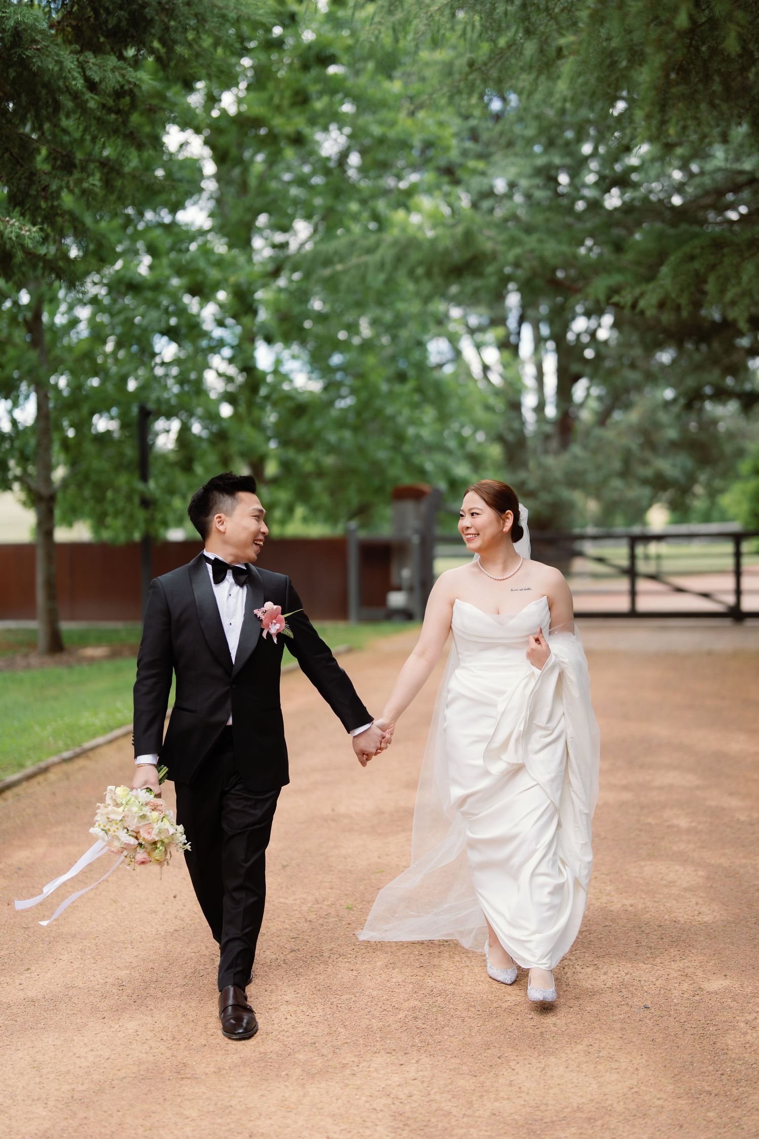 A couple in wedding attire playfully runs and laughs together on a dirt path surrounded by green trees.