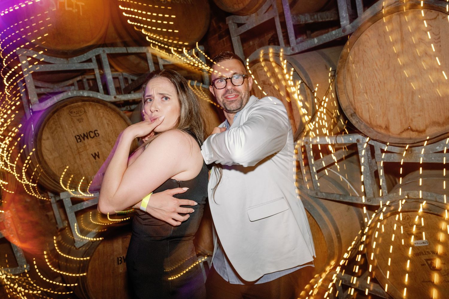 Two people striking playful poses while dancing in front of illuminated wooden barrels at an event venue.