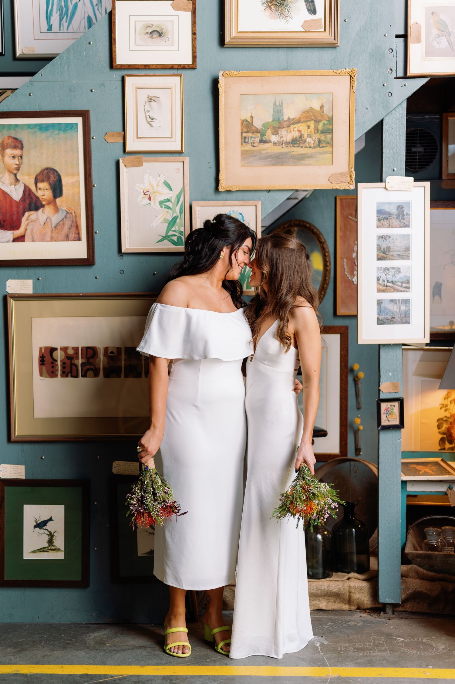 People in white dresses examine artwork displayed on turquoise walls in a gallery setting.