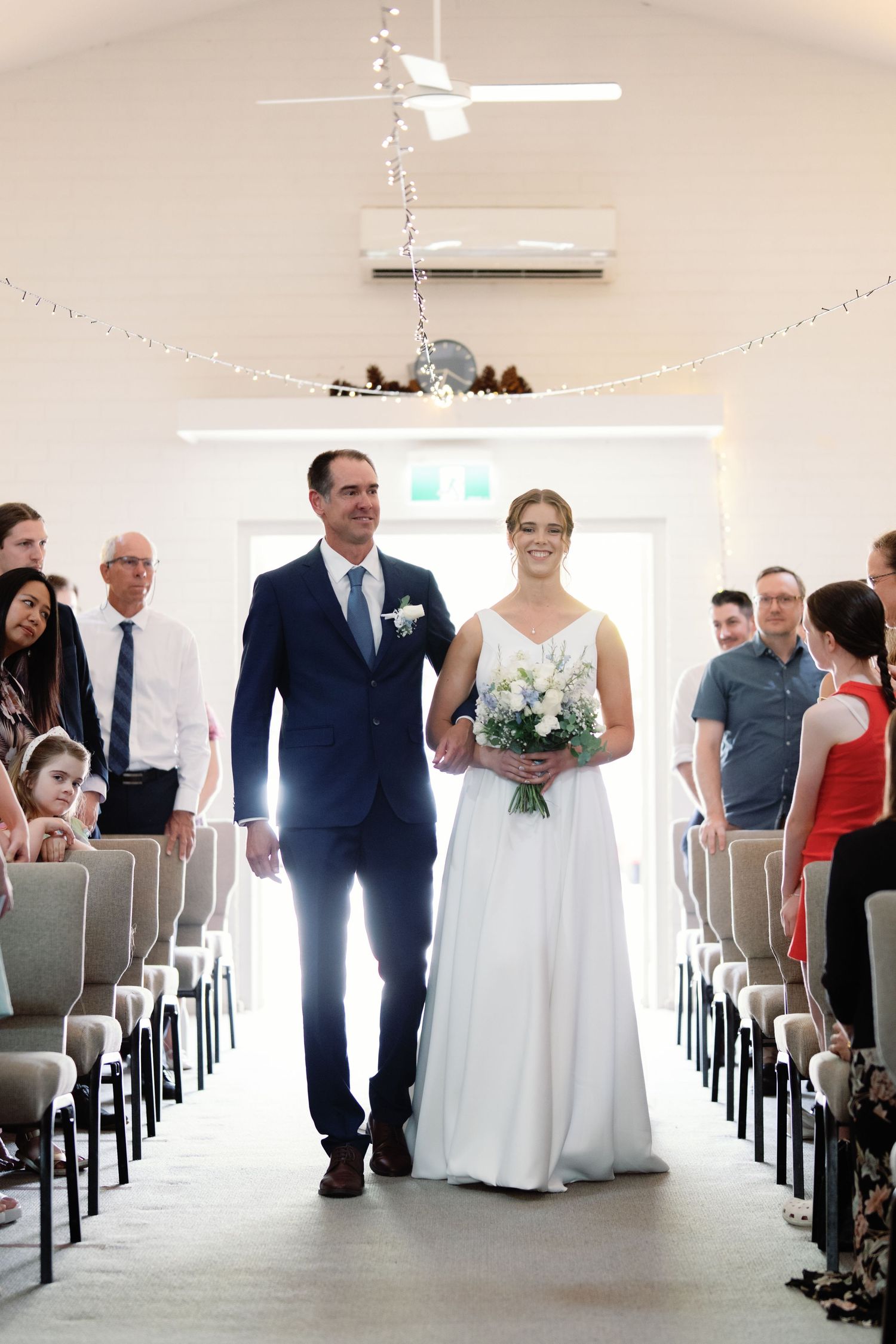 Indoor wedding ceremony setup with white chairs and string lights in bright modern venue.
