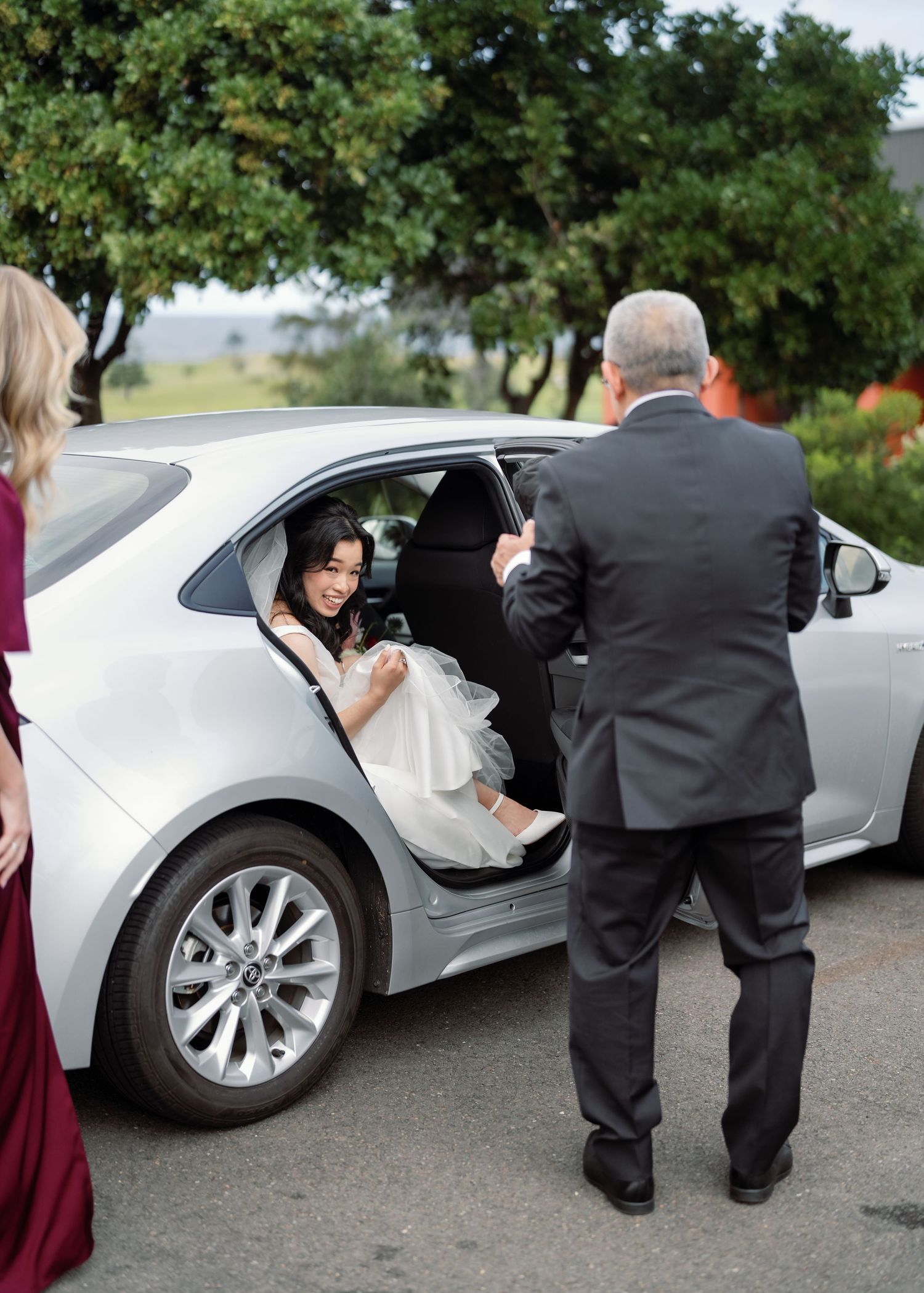 A silver luxury car is parked while formally dressed attendants assist someone entering the vehicle.