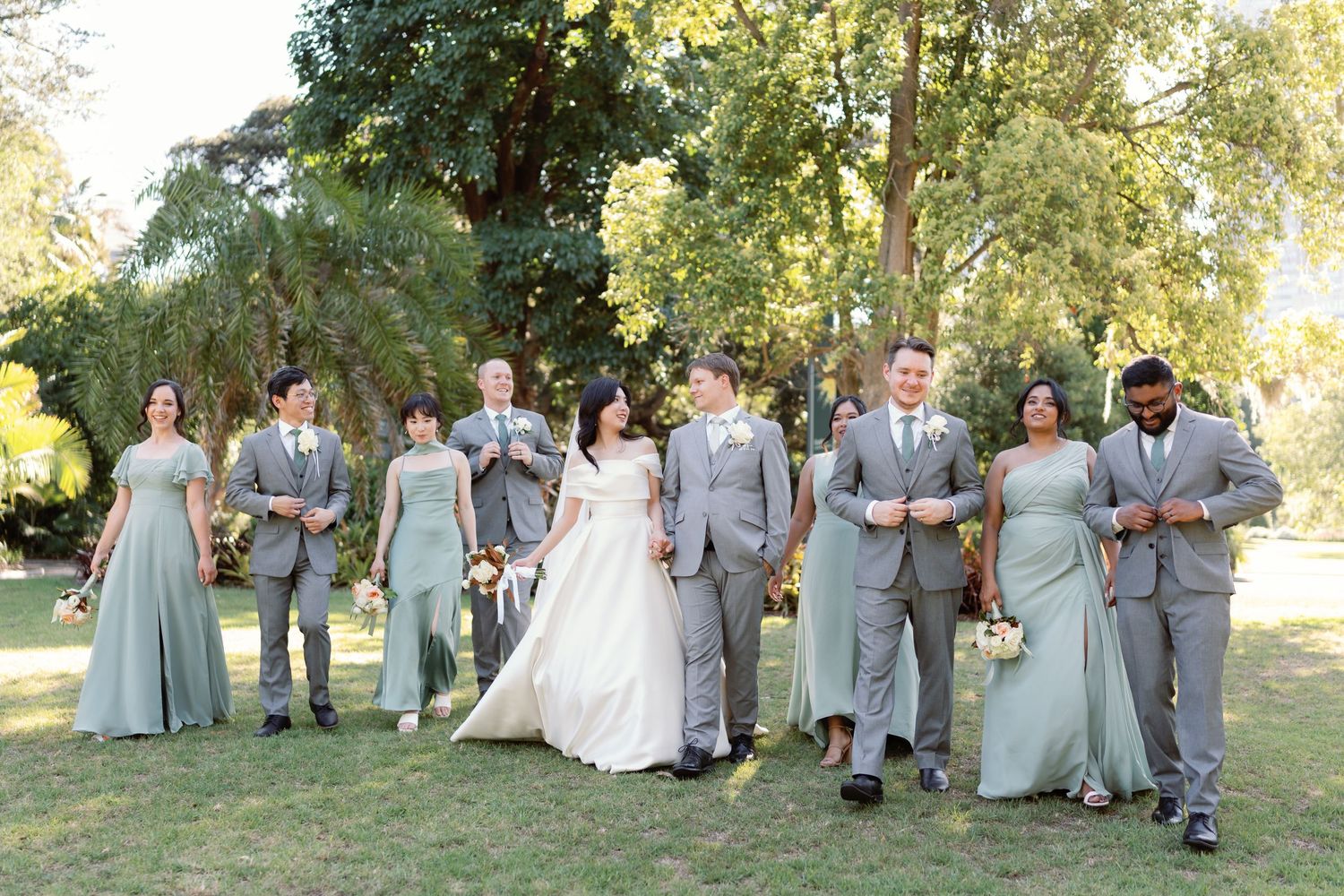 Wedding party in light gray attire poses together on grassy lawn during outdoor summer celebration.