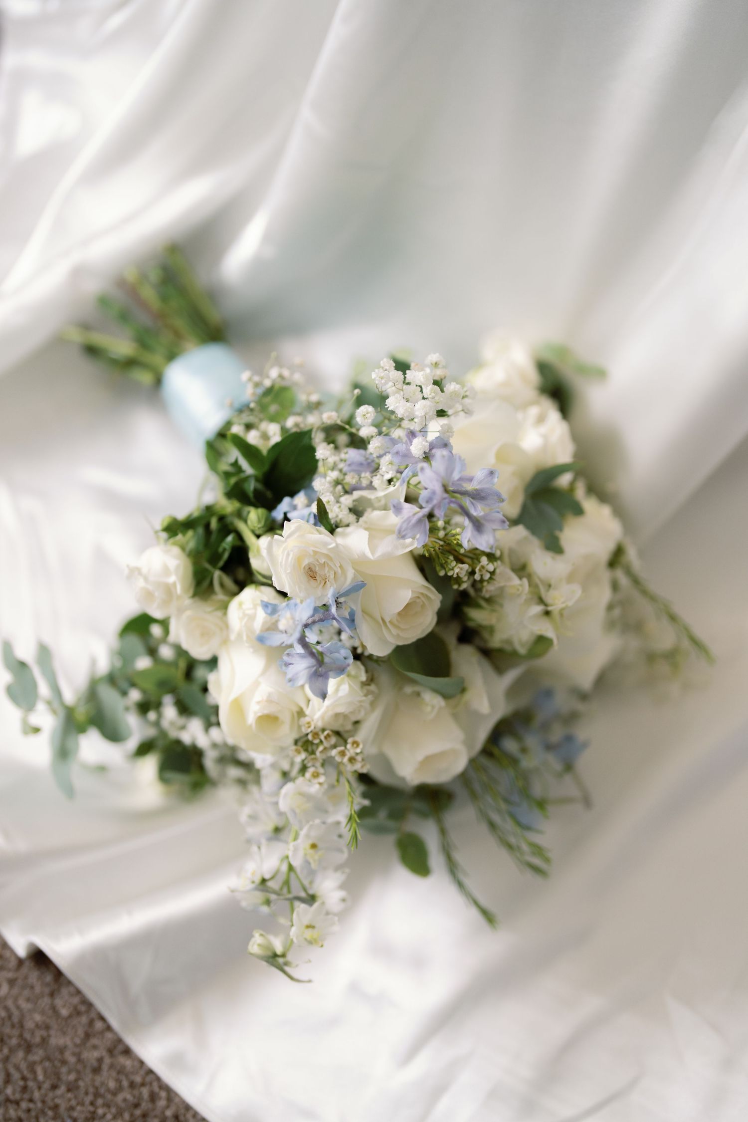 Delicate white and pale blue floral bouquet arrangement with greenery against white fabric background.