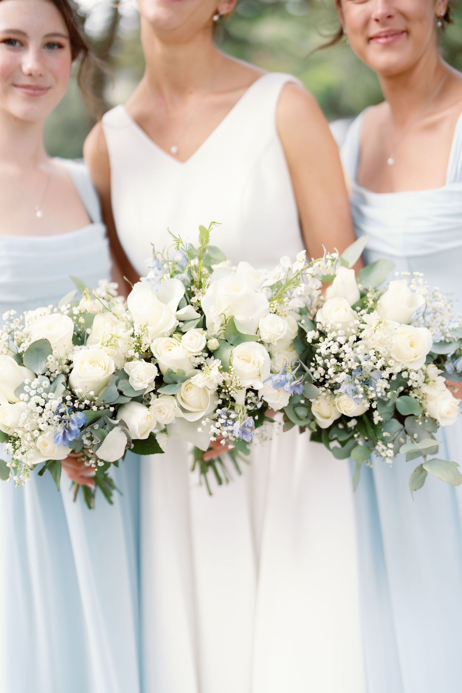 Bridesmaids in light blue dresses hold elegant white rose and greenery bouquets in a wedding party lineup.