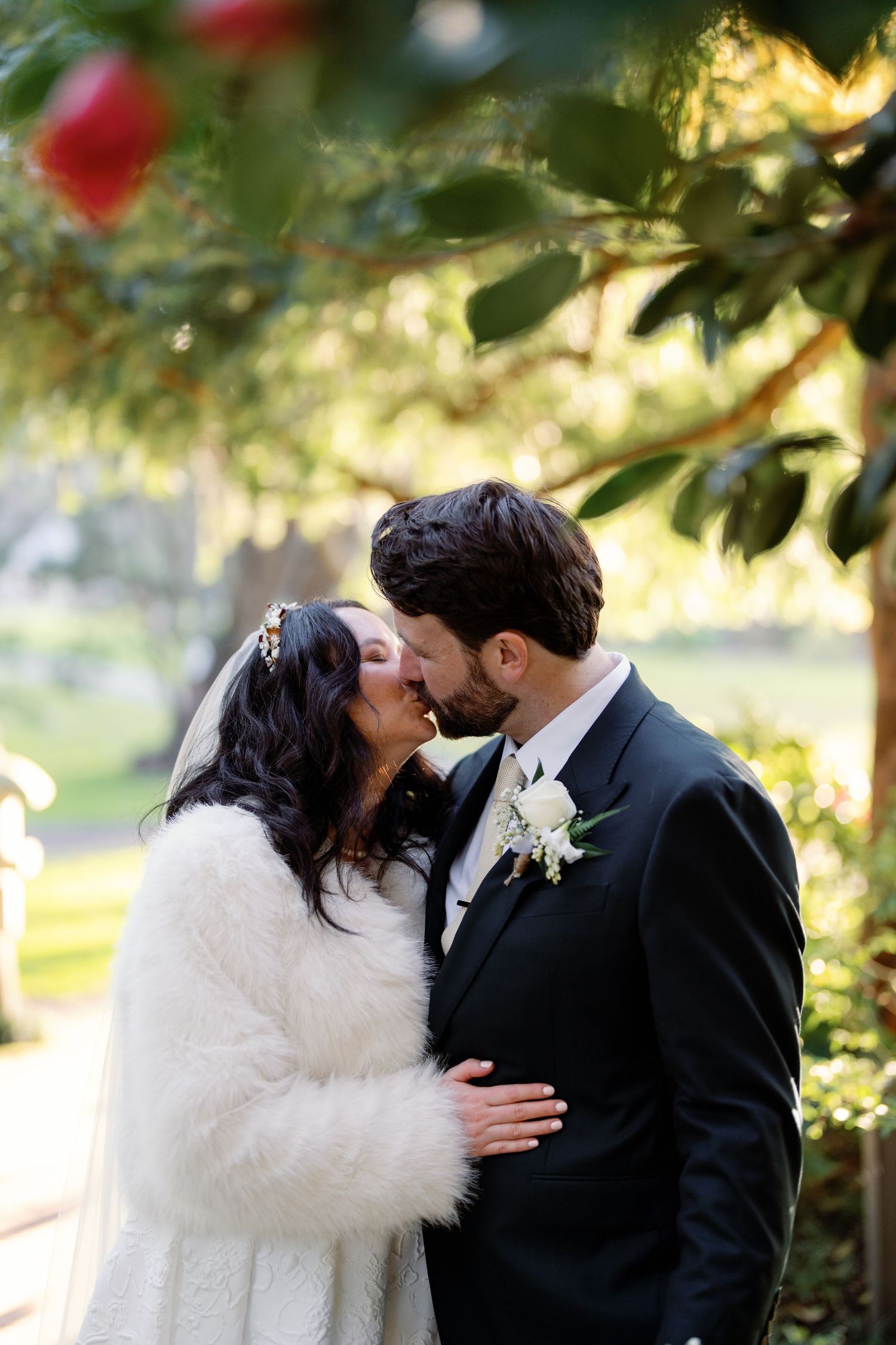 Bride in white fur stole and groom share an intimate kiss surrounded by lush garden greenery.