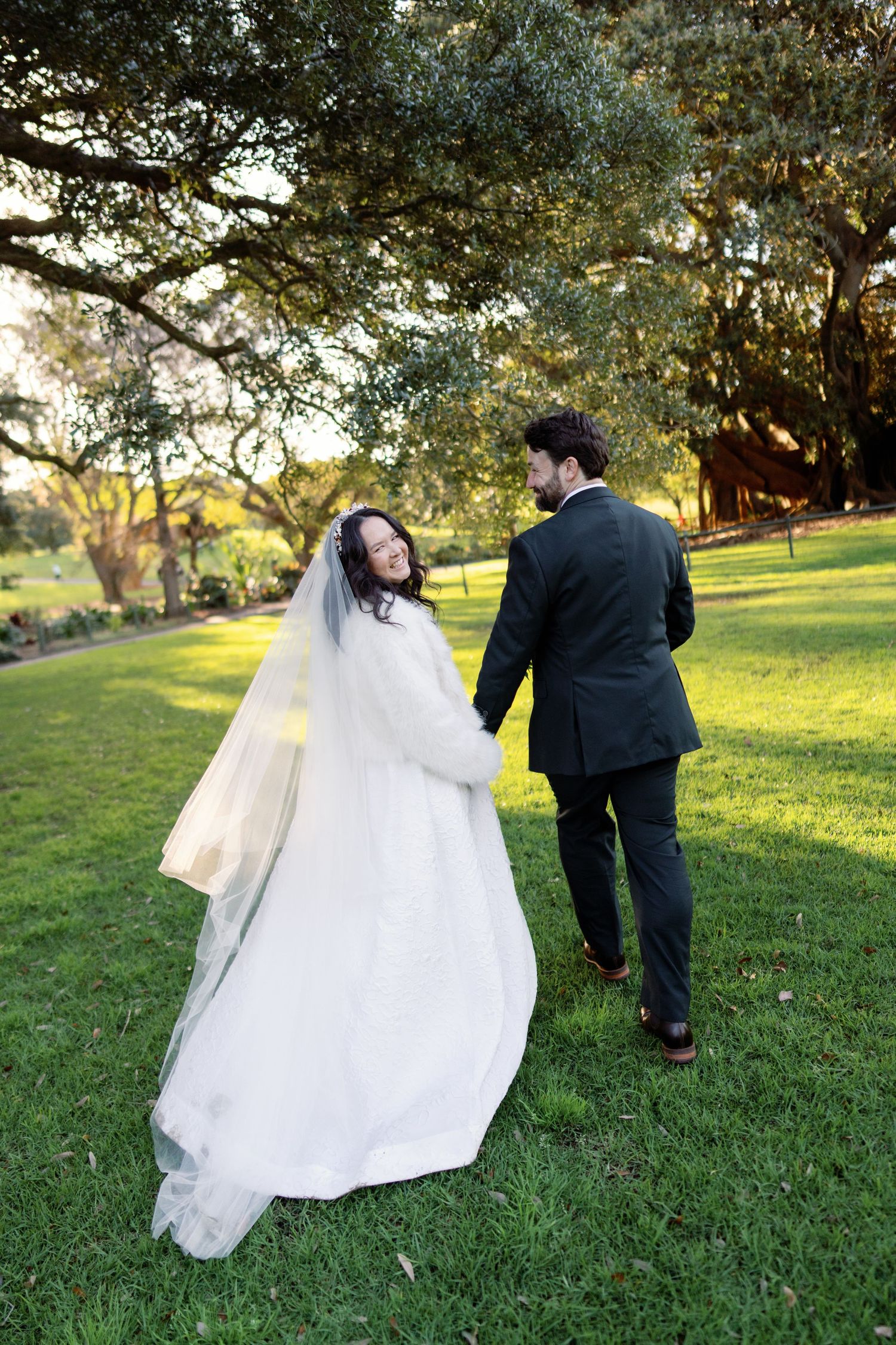 Bride in white gown and veil holds grooms hand walking through sunlit park.