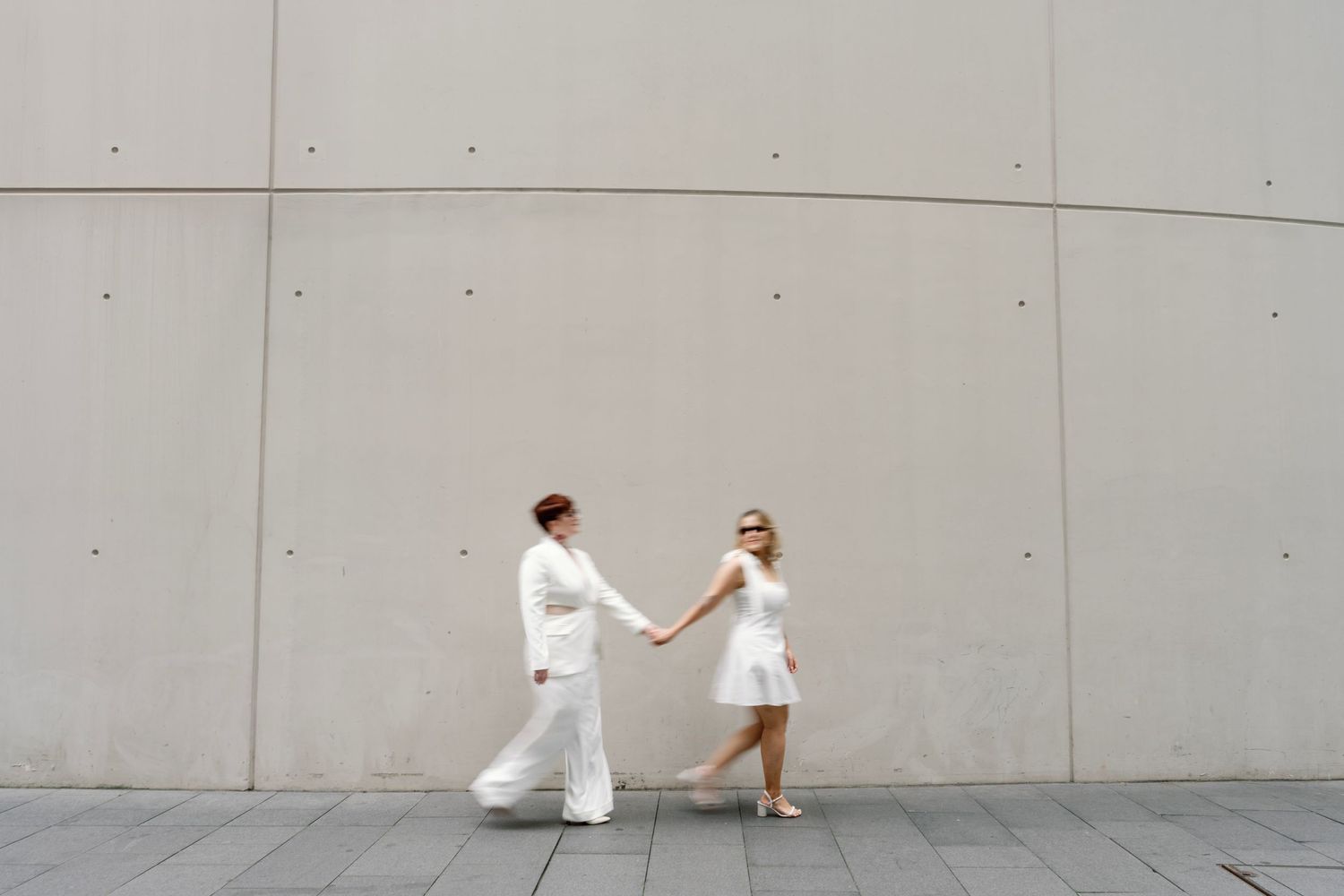 Two figures in white outfits hold hands against a minimalist grey wall backdrop.