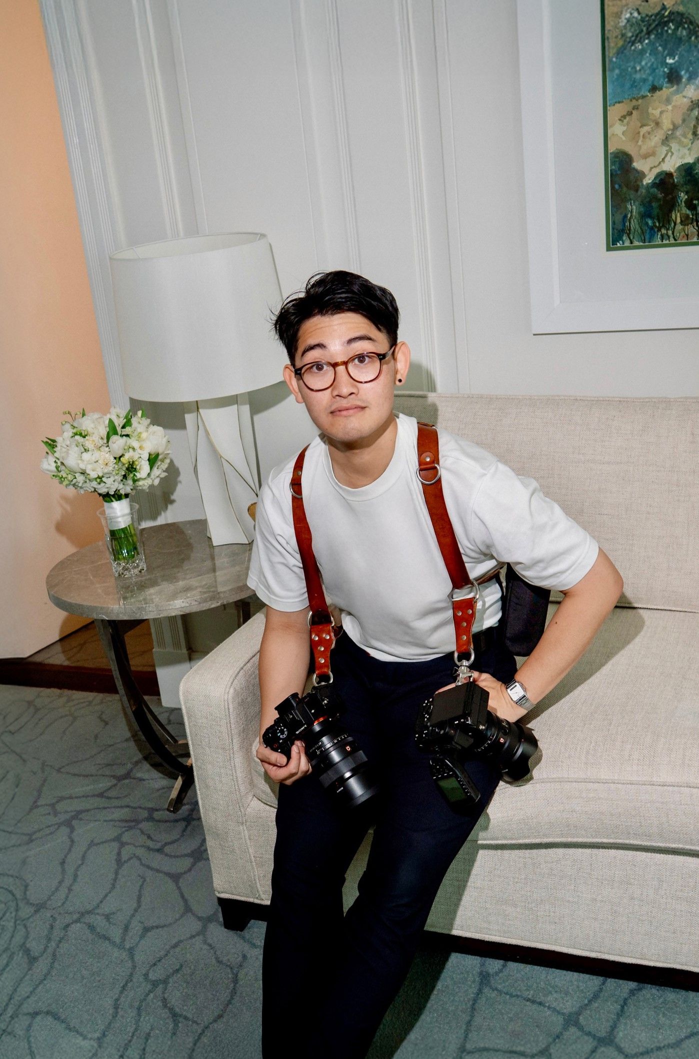 A photographer with dual camera setup poses in a living room with landscape art.