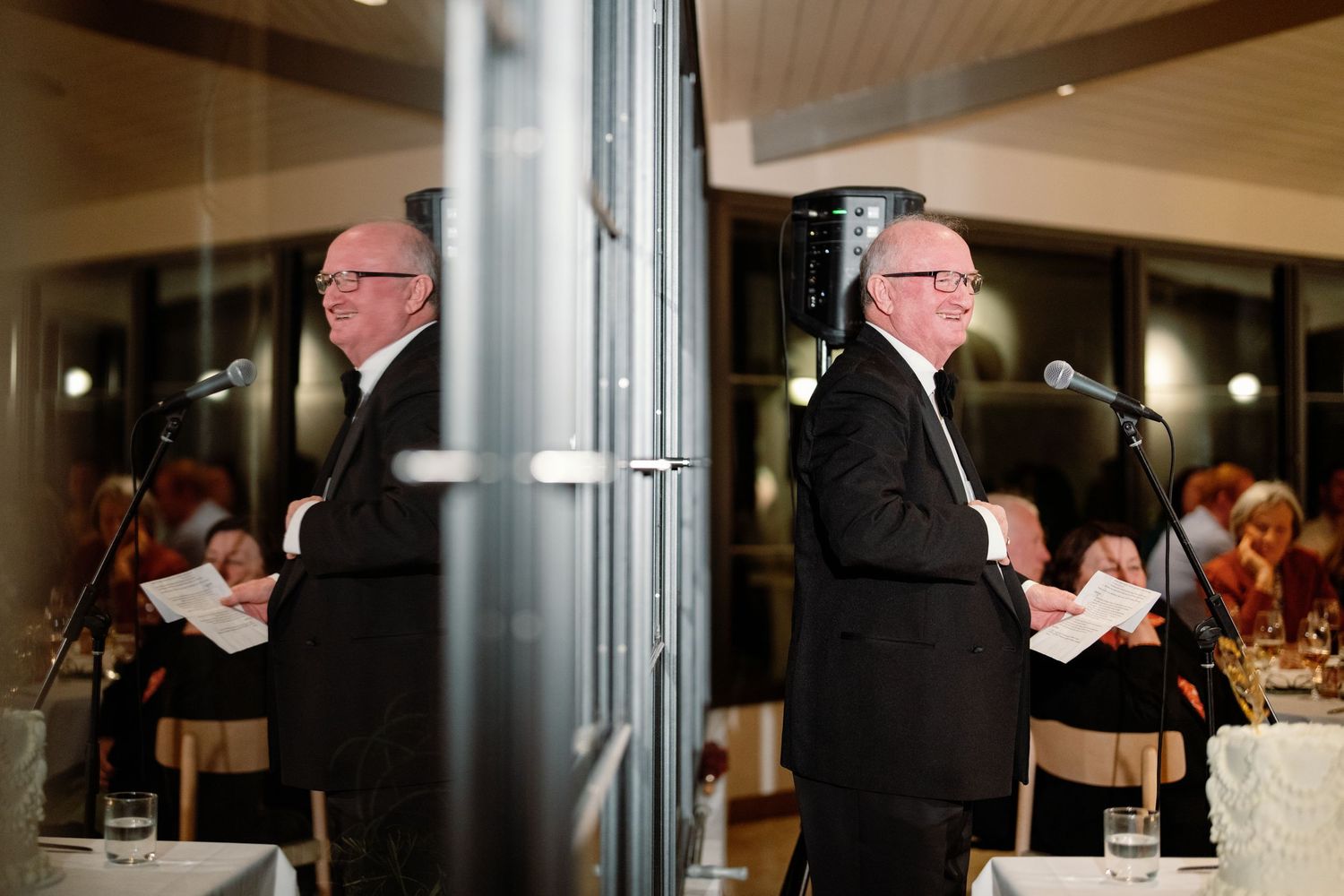 Man in black suit giving a speech at a podium reflected in a mirror at a formal dinner event.