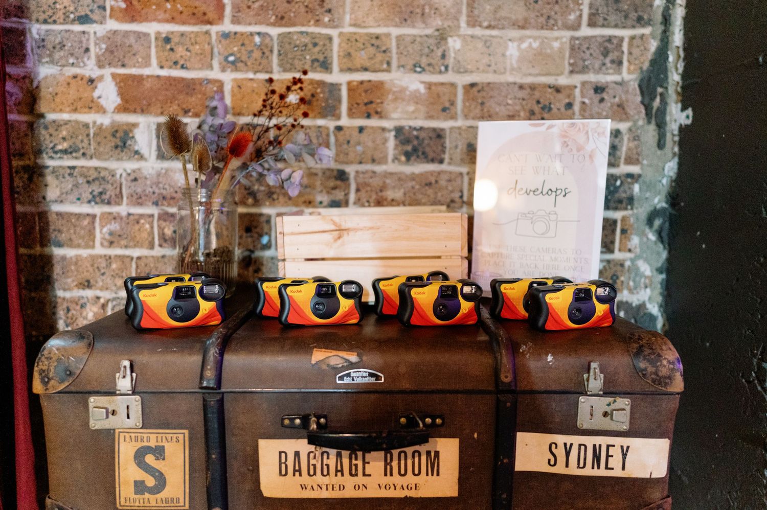 Vintage luggage and wooden crates stacked against exposed brick wall with 'Sydney' label visible.