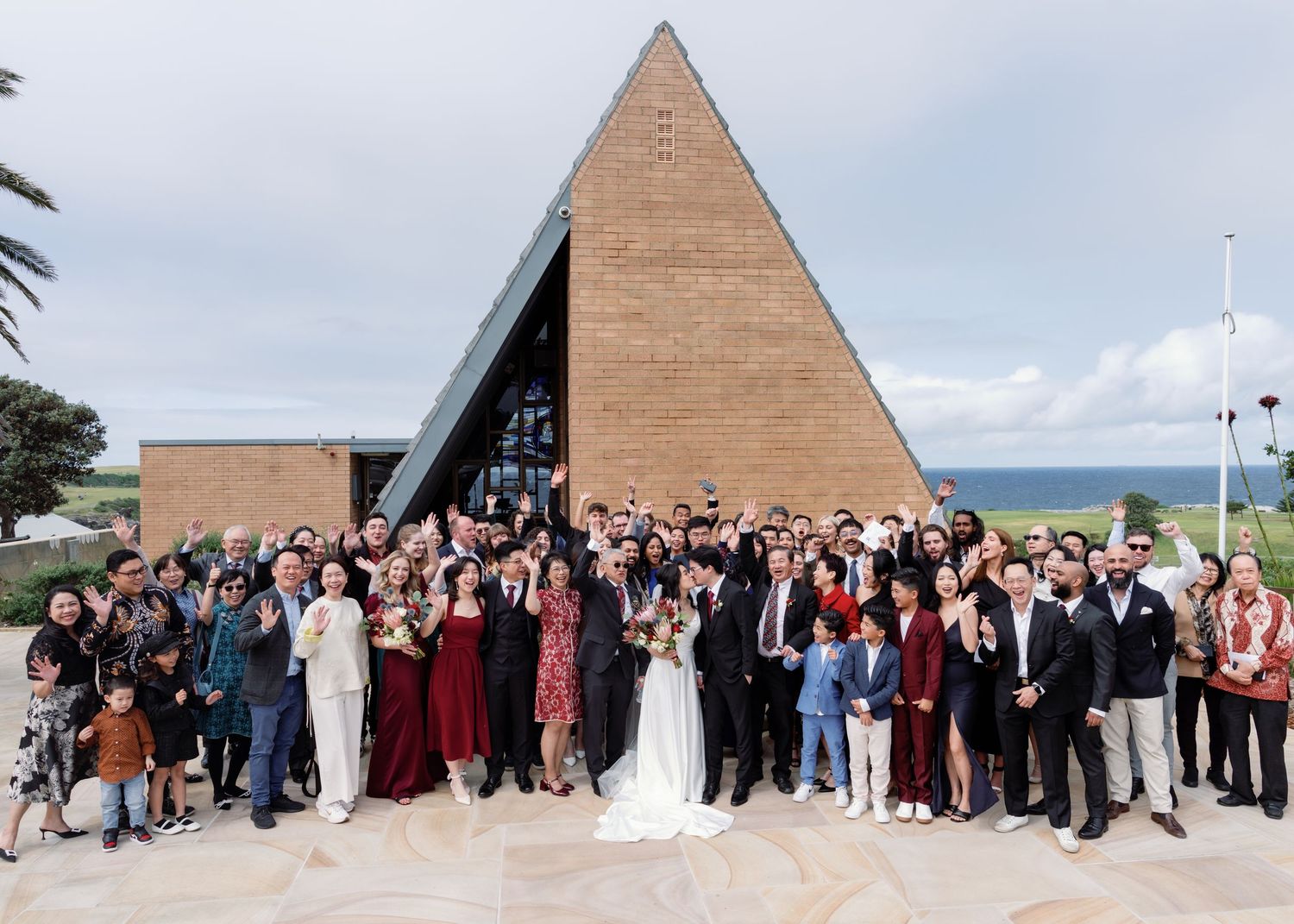 Large group of wedding guests pose for panoramic photo in front of triangular wooden building structure.
