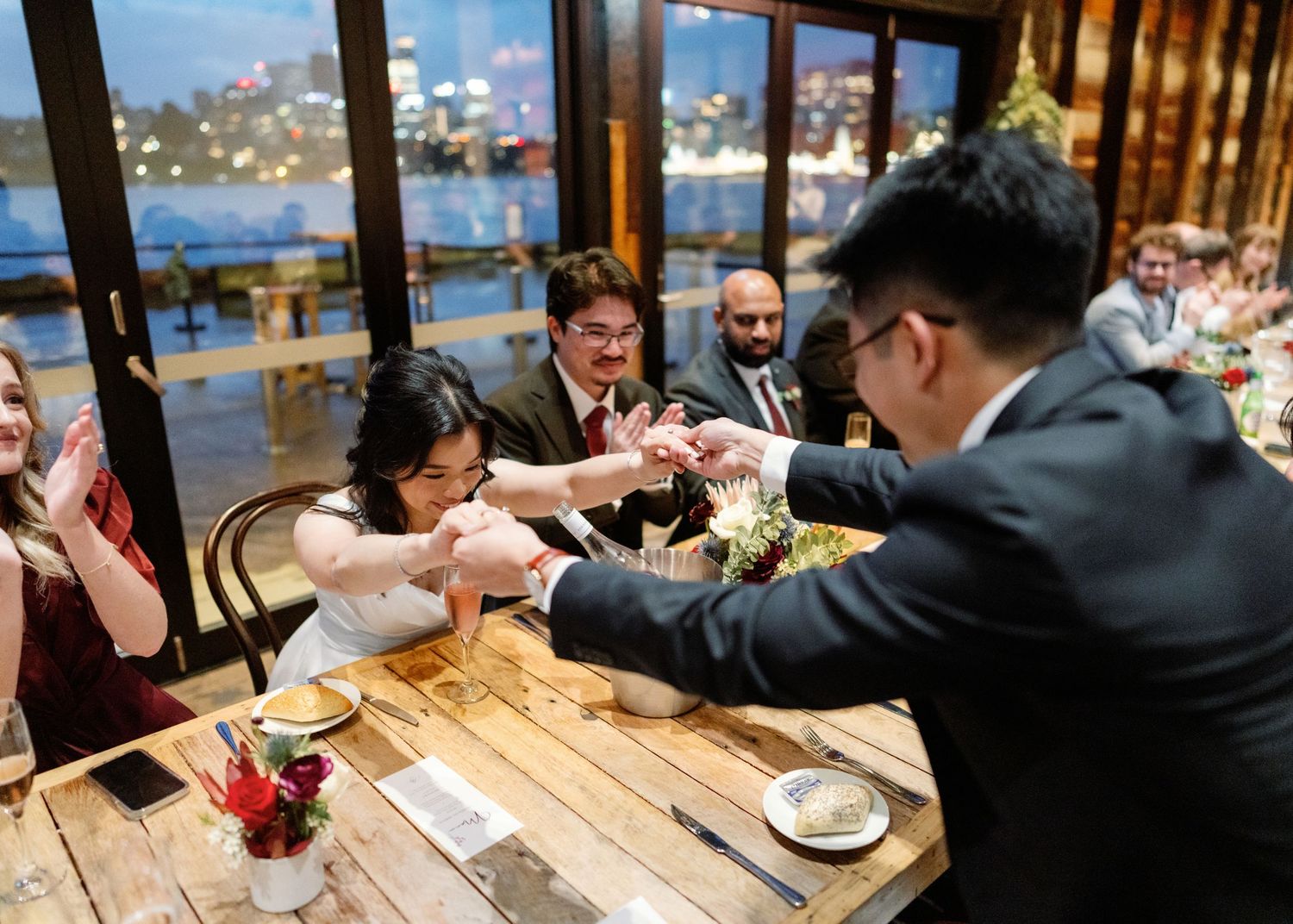 Wedding reception guests dining at rustic wooden tables with waterfront view.