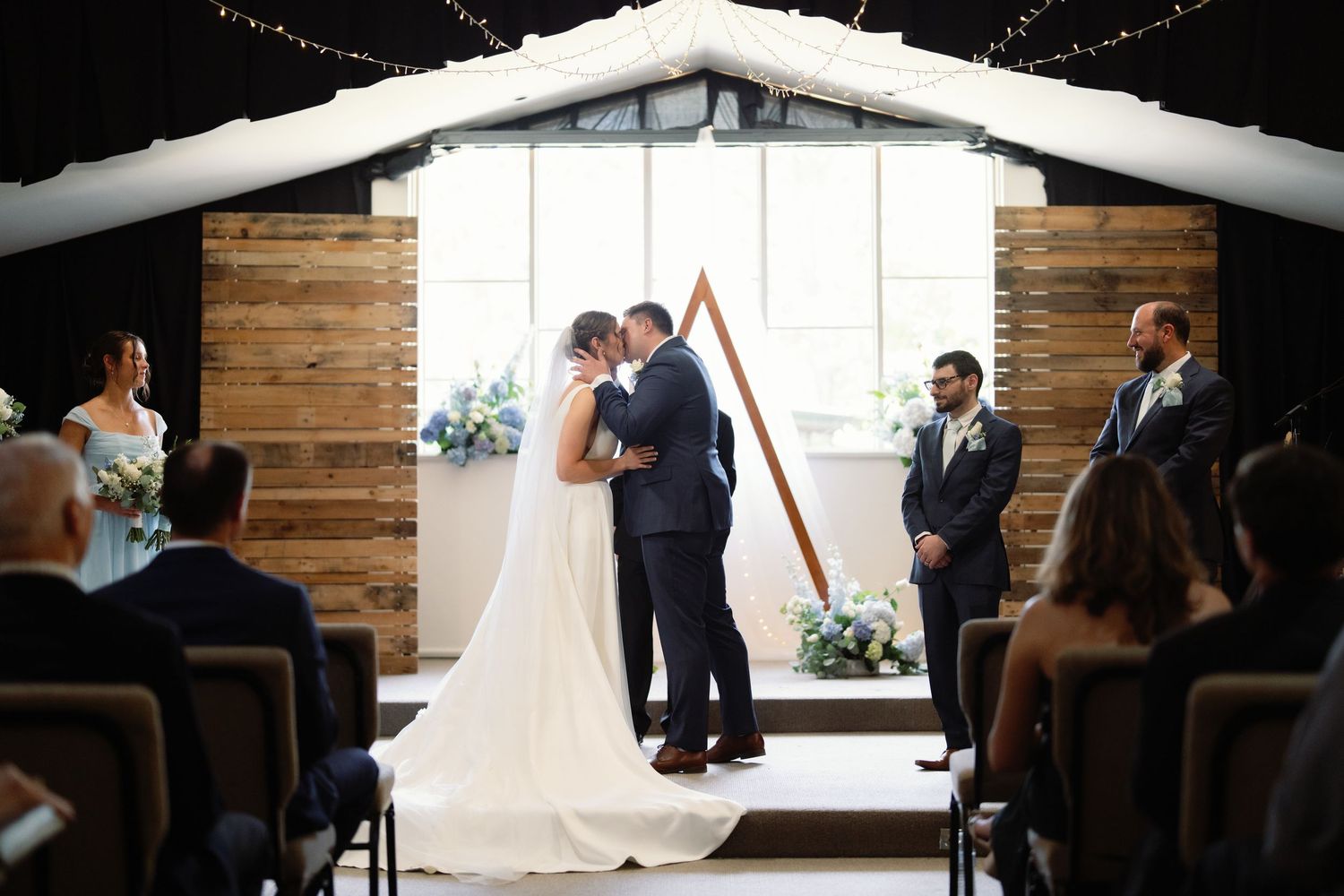Wedding ceremony taking place in a rustic indoor venue with wooden walls and rows of guest seating.