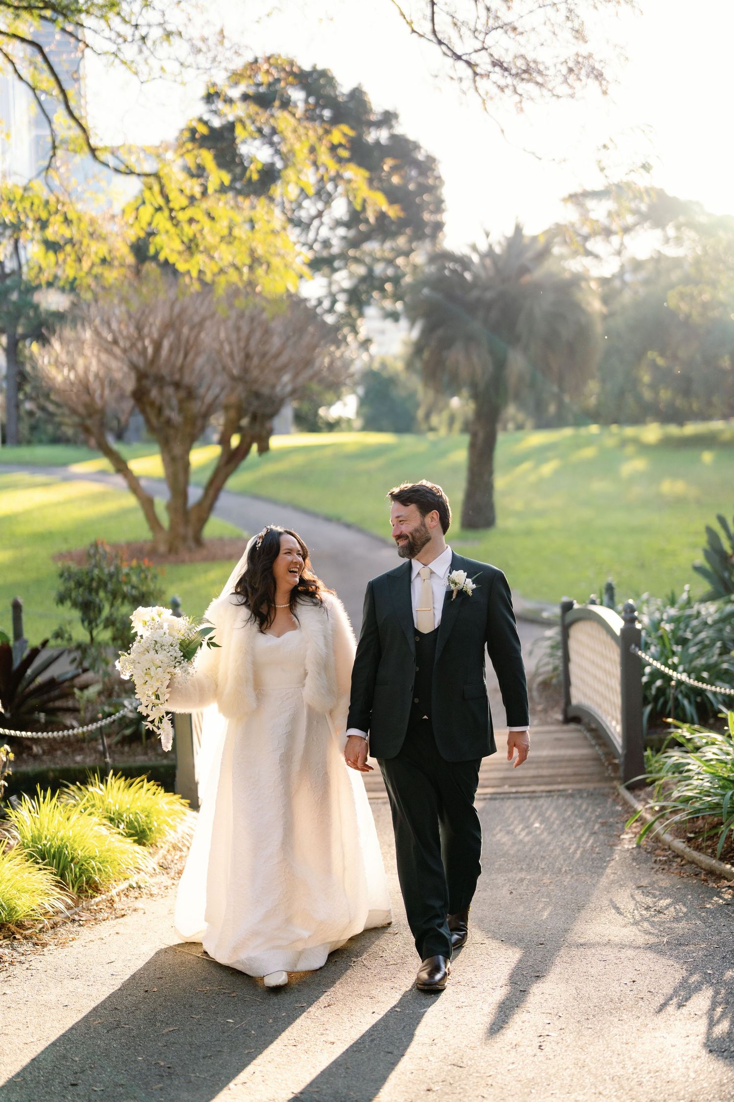 Bride and groom walk hand-in-hand through a sunlit garden path, bride wearing a white gown and fur wrap.