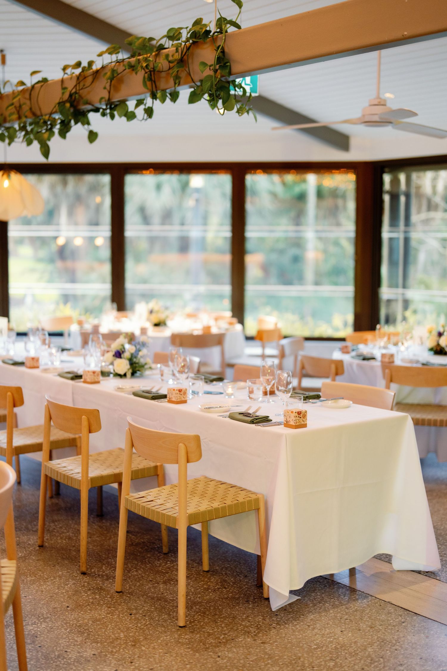 Elegant wedding reception venue with long white linen tables, wooden chairs, candle centerpieces, and greenery overhead in a bright room.