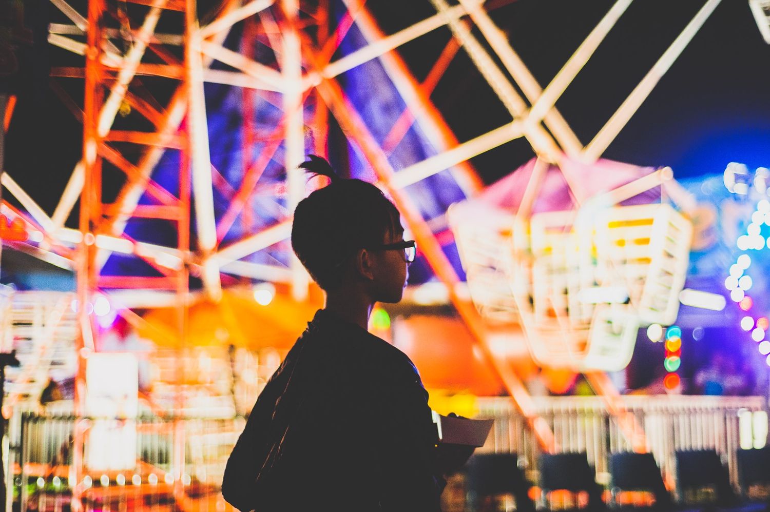 A silhouette of a person looking at illuminated carnival rides at night.