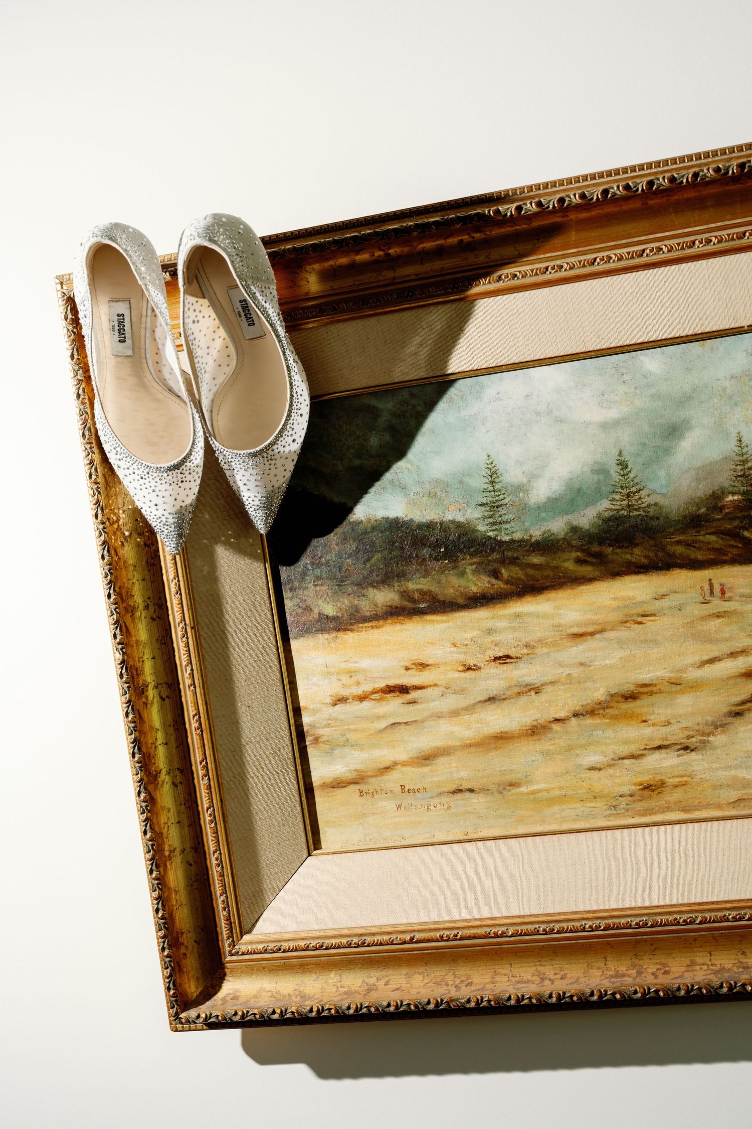 A framed landscape painting with a pair of white shoes displayed on top of the frame.