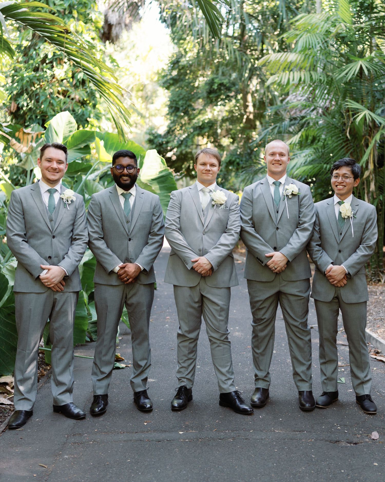 Groomsmen in matching gray suits pose in a line against a lush garden backdrop.