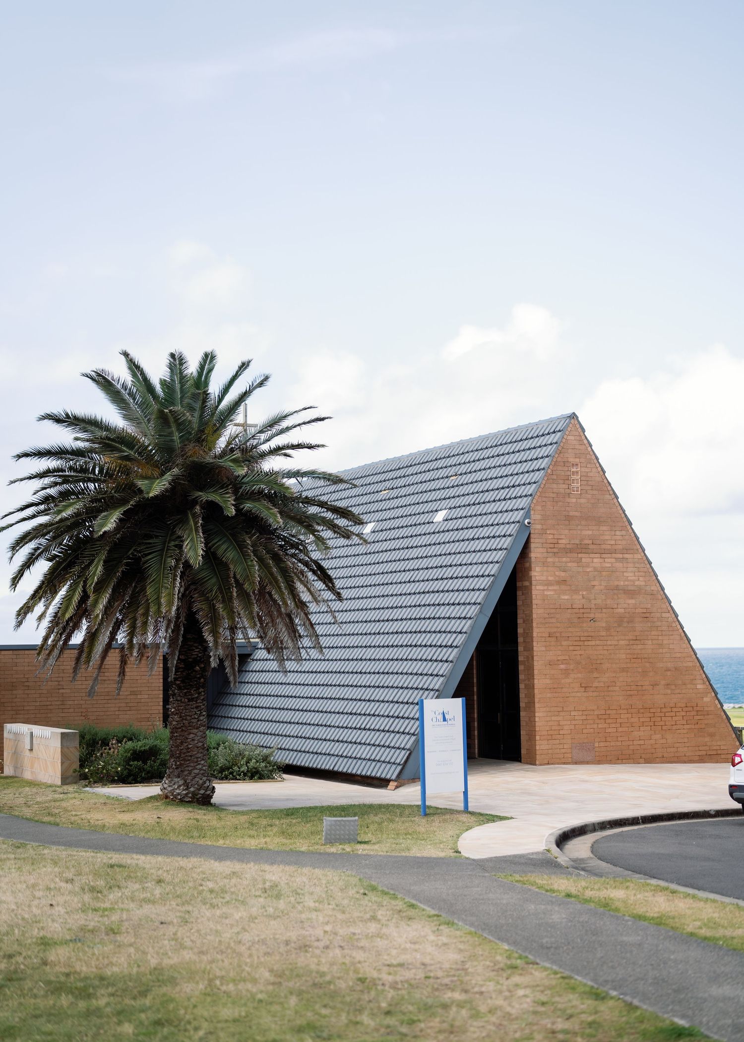 Modern triangular buildings with slanted roofs and a palm tree stand against a cloudy sky at a coastal location.