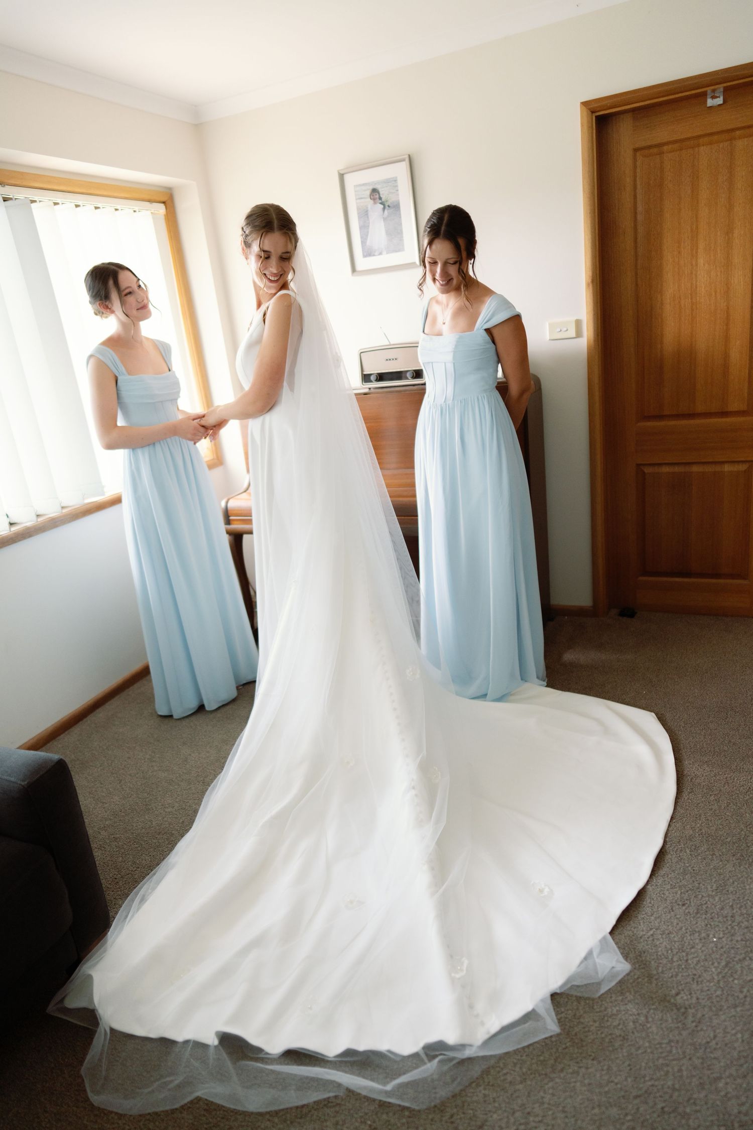 Bridesmaids in light blue dresses helping with a flowing white wedding gown in a sunlit room.