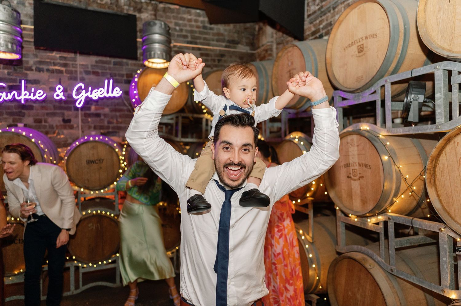Festive celebration with dancing guests in a barrel room decorated with string lights.
