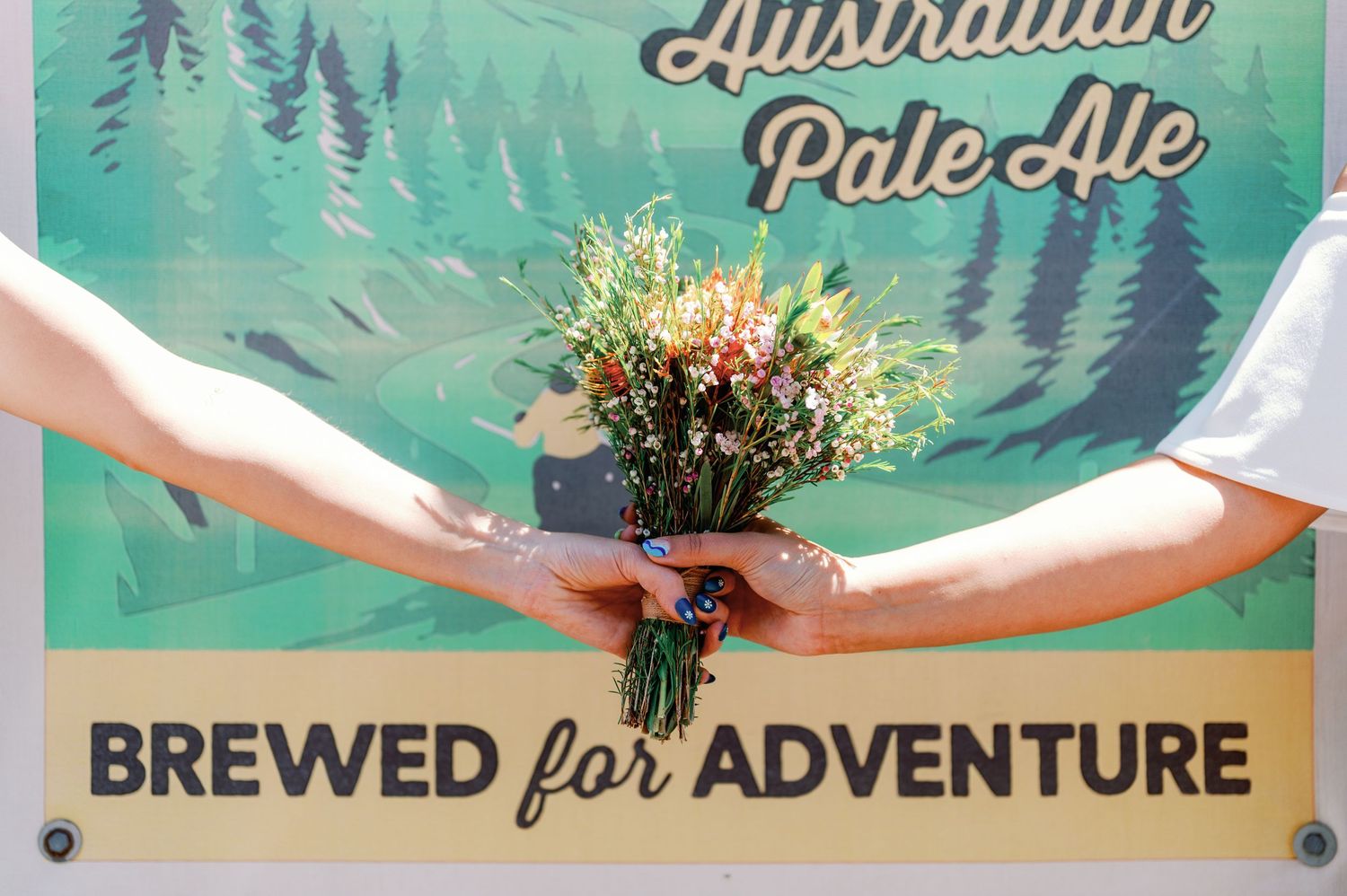 A bouquet of wildflowers held between hands against a brewery sign with teal forest artwork in the background.