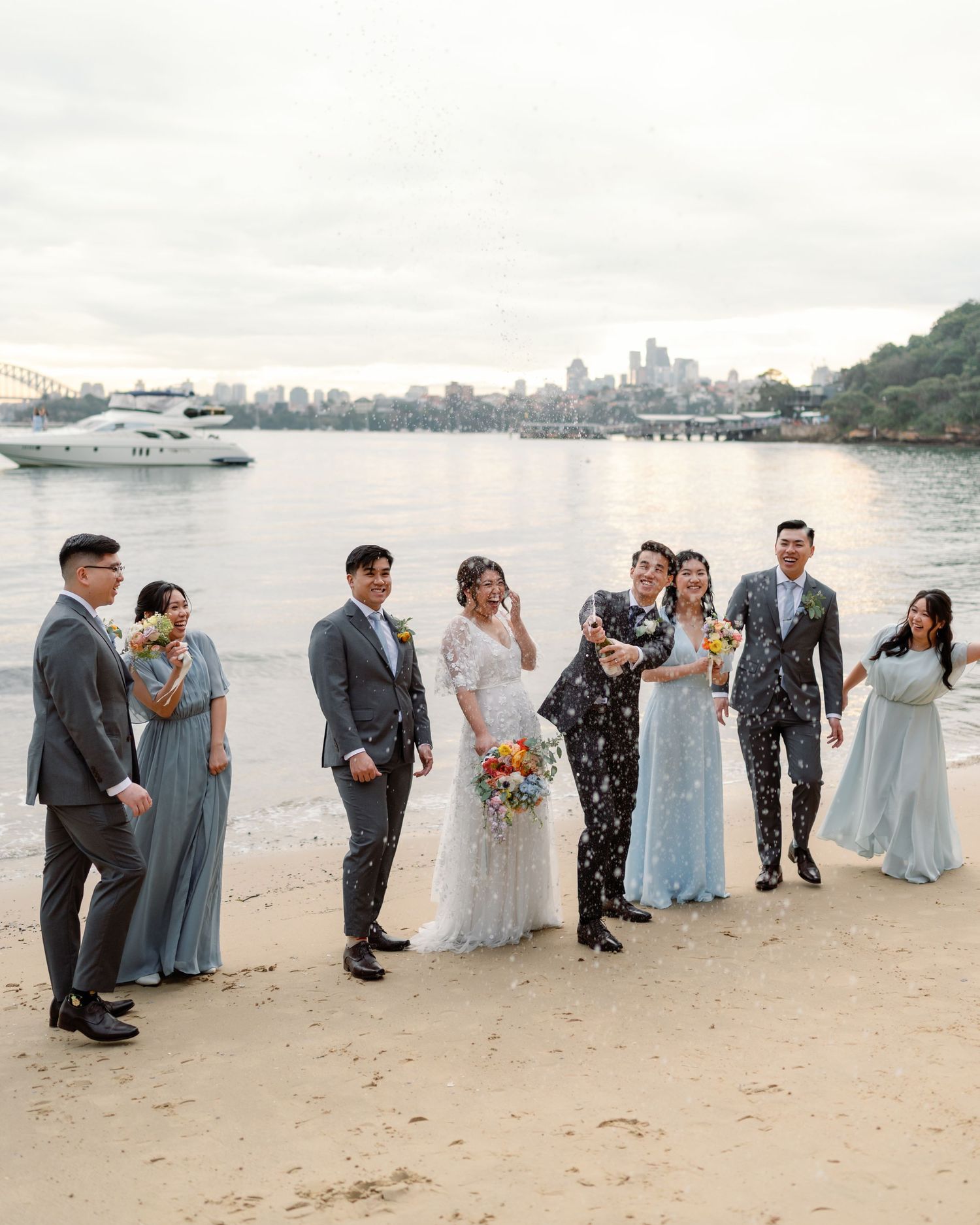 A wedding party celebrating on a beach with boats and city skyline in the background.