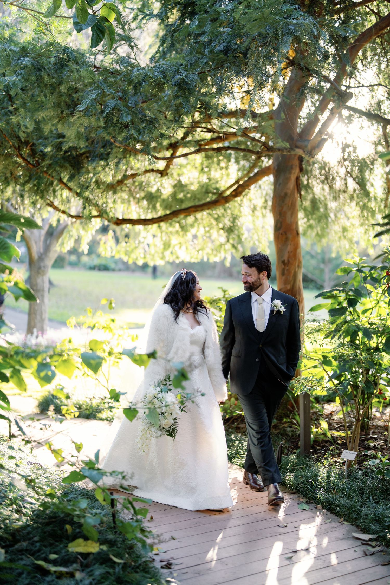 Bride and groom walk hand-in-hand on a wooden path surrounded by lush greenery in golden sunlight.