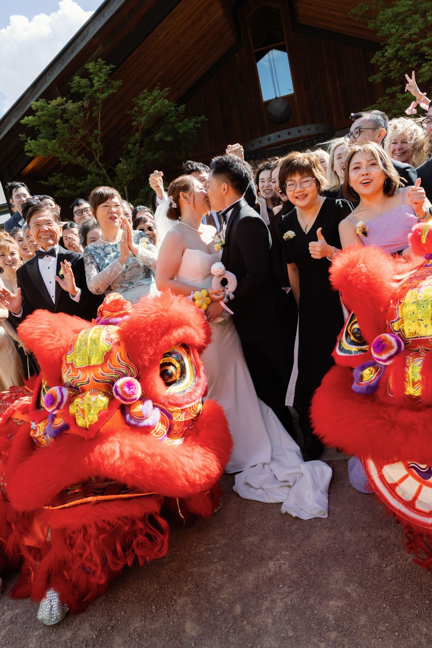 Traditional Chinese lion dance performers in bright red costumes celebrate at a wedding ceremony outside a wooden building.