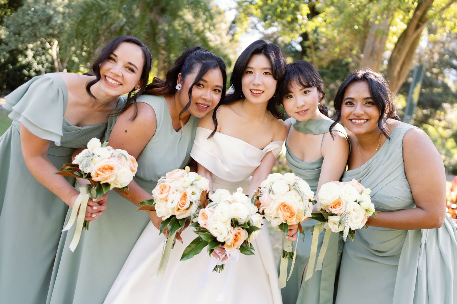 Bridal party in sage green dresses poses outdoors with bouquets of white and peach flowers.