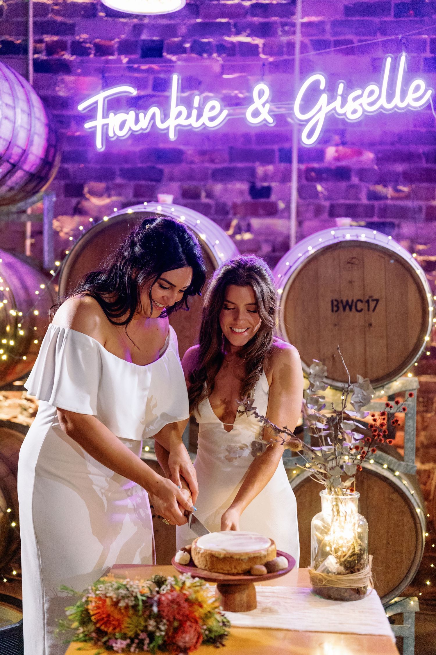 Two people in white dresses cut a wedding cake together under purple lighting with wooden barrels in the background.