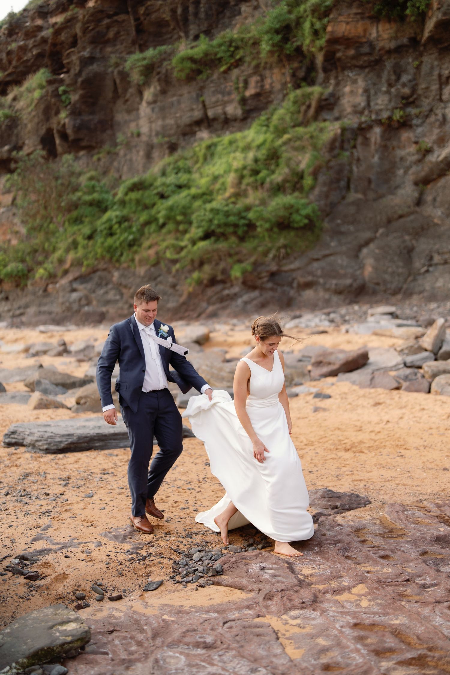 A couple in wedding attire playfully interacts on a rocky beach with cliffs in the background.