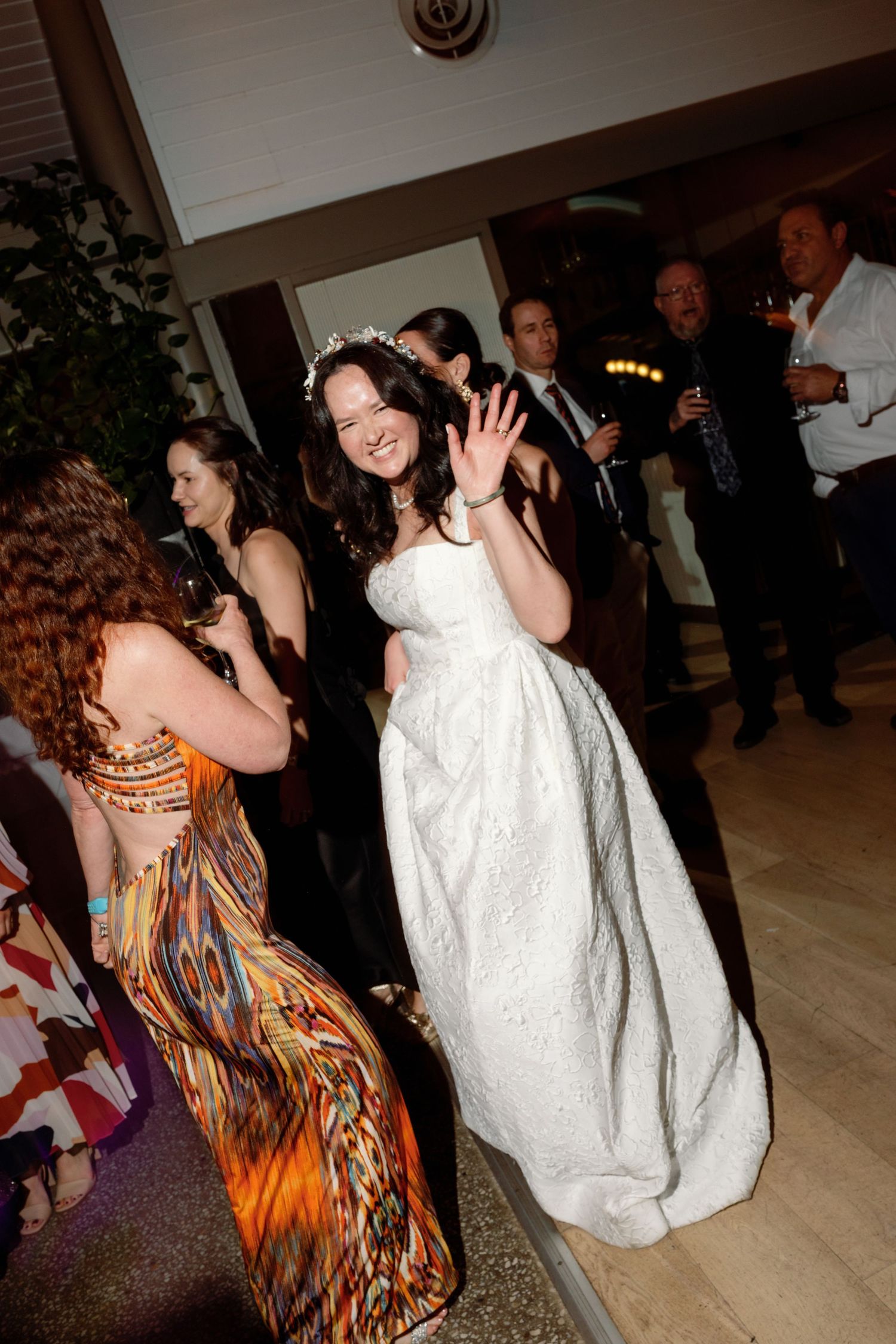 Bride in white ballgown waving and dancing with guests at indoor wedding reception celebration.