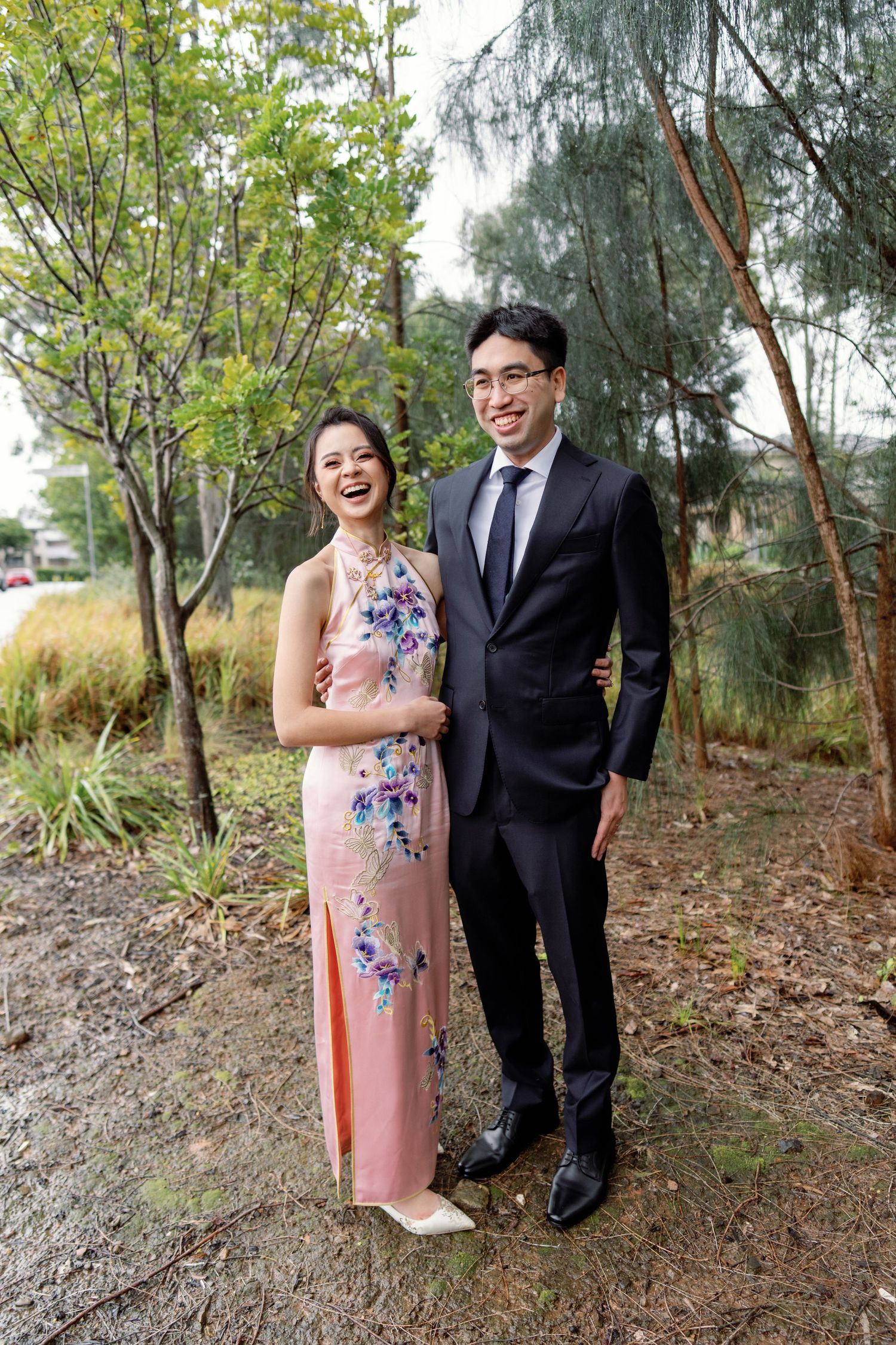 A couple poses together outdoors in formal attire with trees and natural scenery in the background.