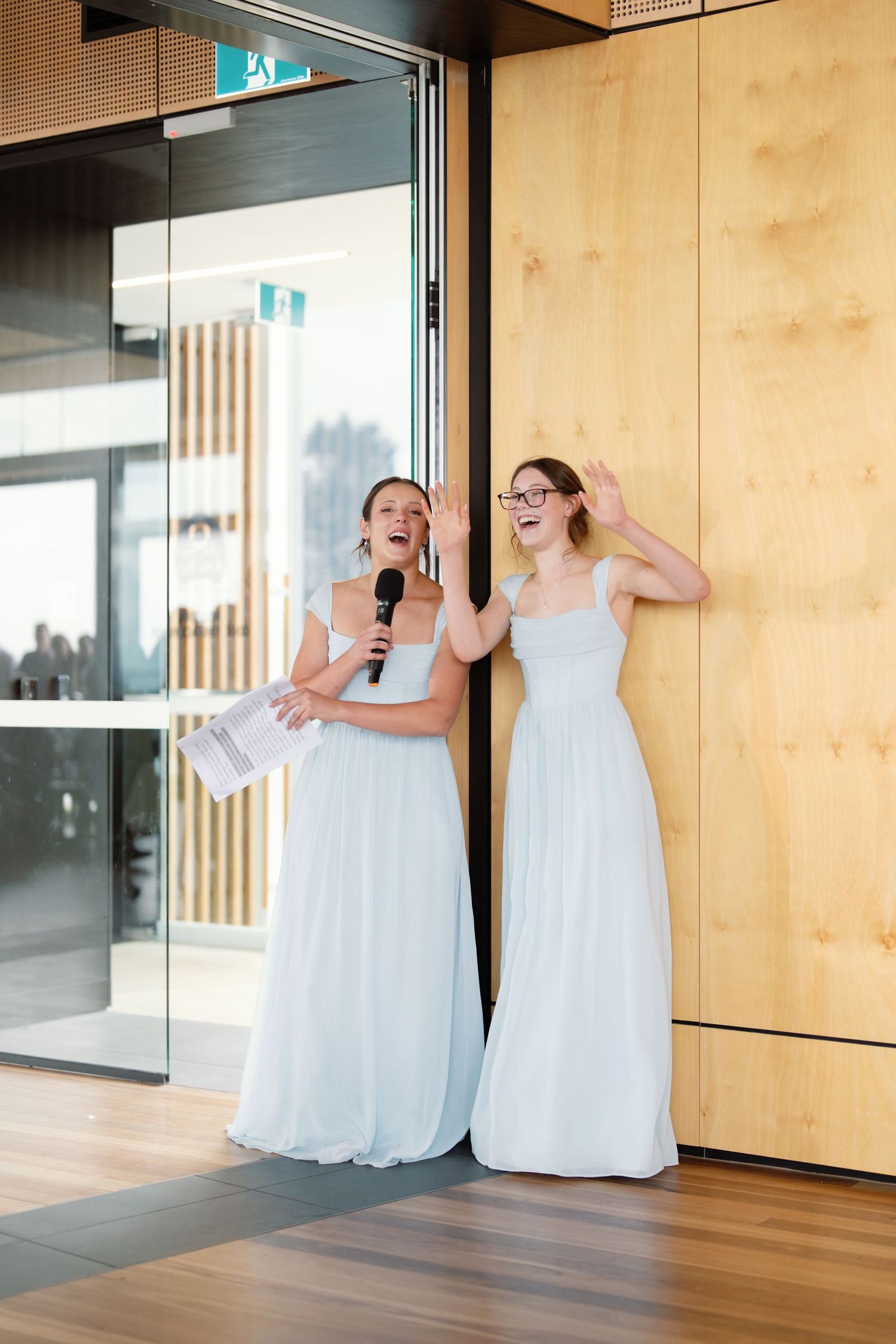 Bridesmaids in white dresses share a laughing moment in a modern wood-paneled hallway.