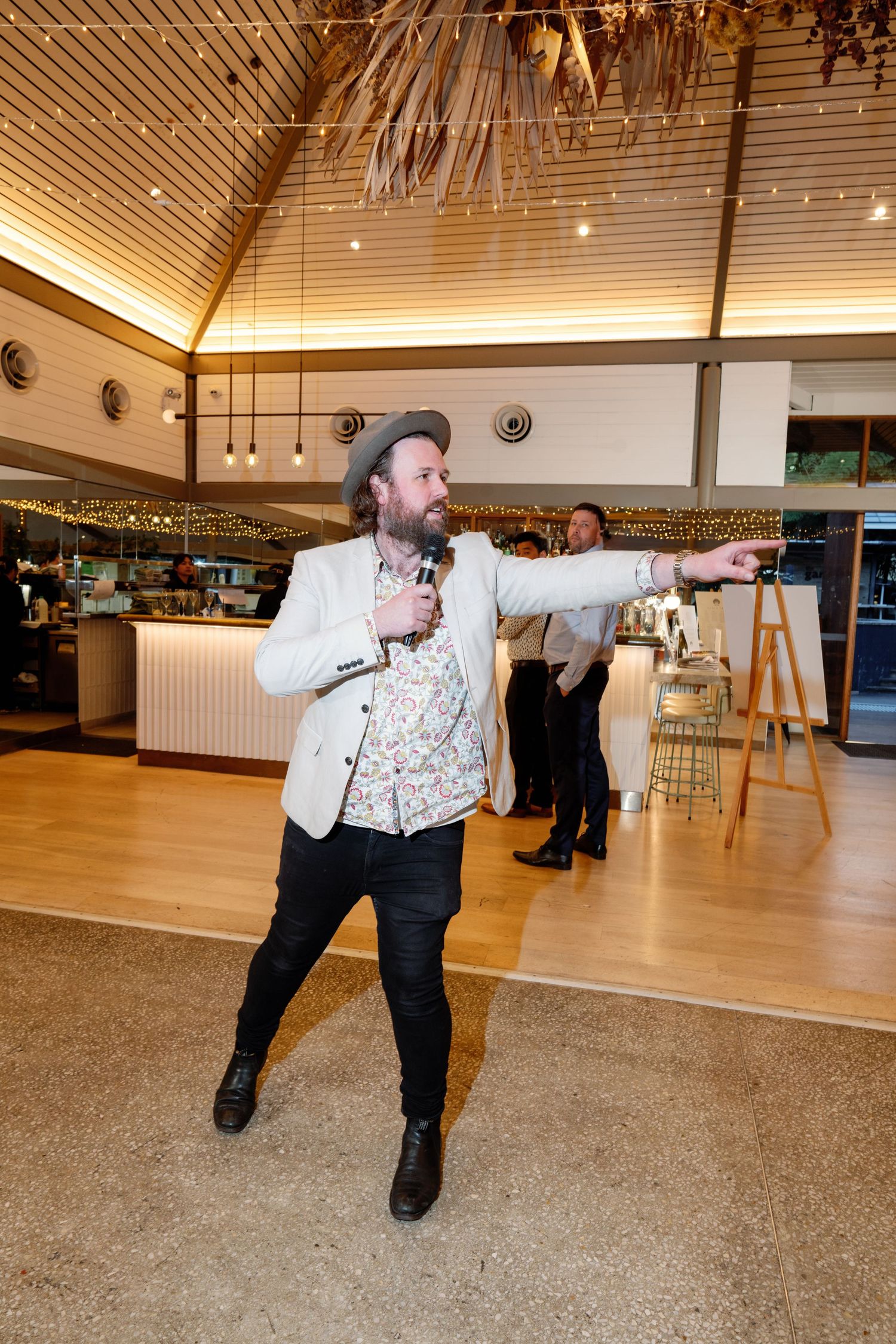 Energetic MC in hat and floral shirt performs with microphone at wedding reception.