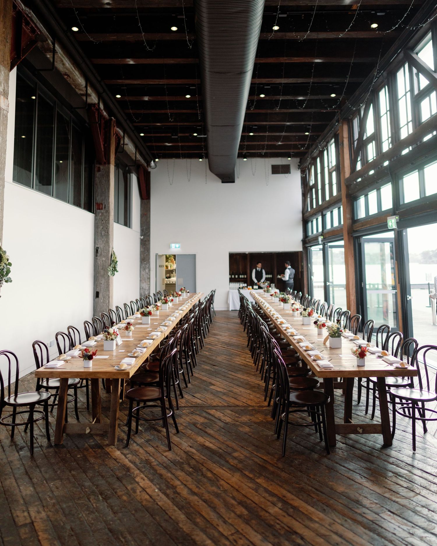 Long wooden dining tables with bentwood chairs set up in an industrial-style wedding reception venue.