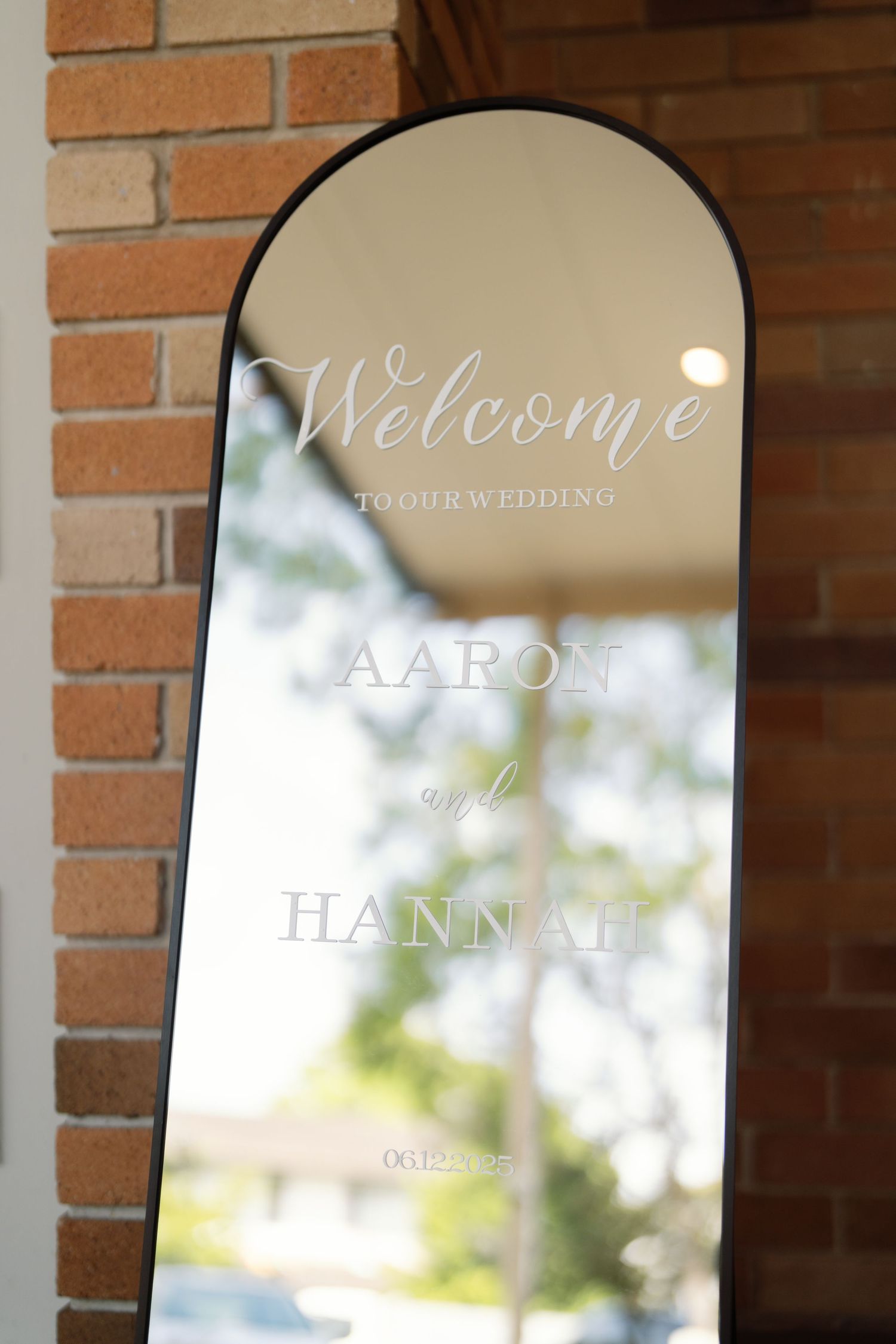Decorative welcome sign with elegant script lettering on arched mirror against brick wall.