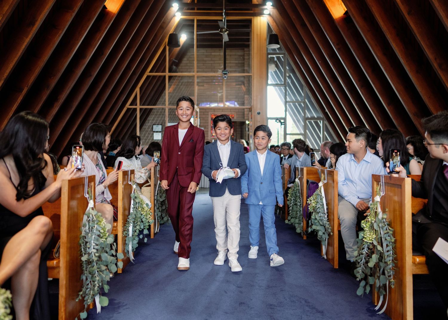 Wedding party walking down a rustic wooden aisle decorated with greenery and warm lighting at an indoor venue.