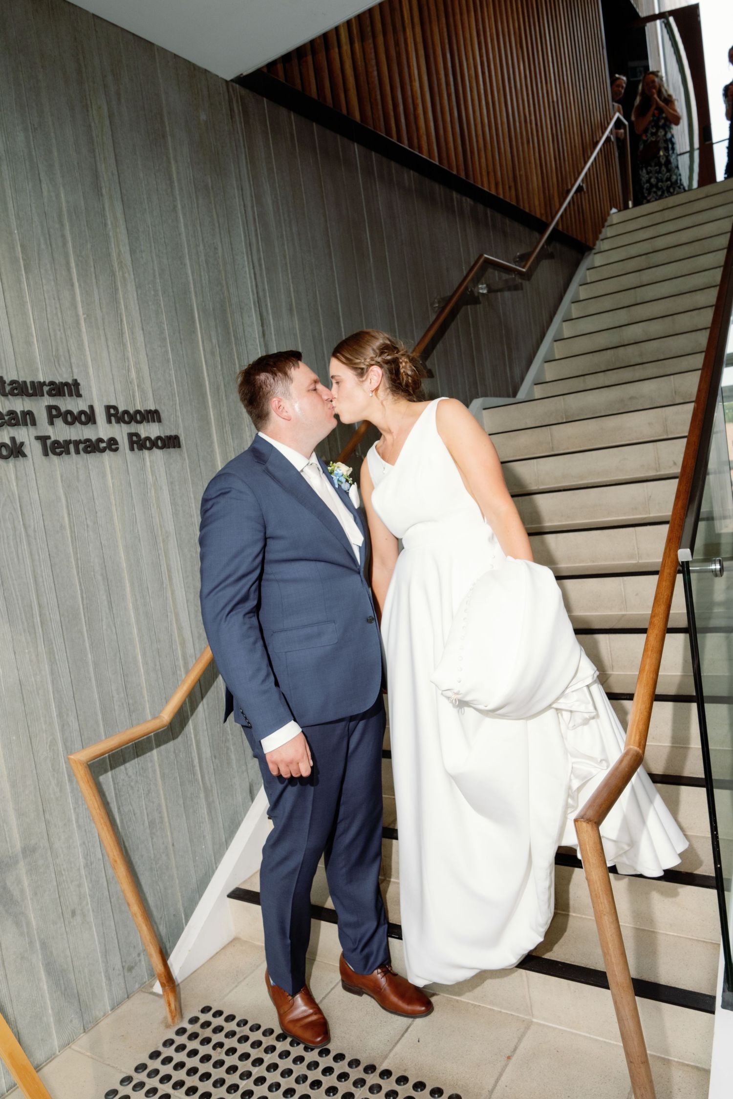 Wedding couple ascends modern concrete staircase with wooden handrails in an indoor venue.