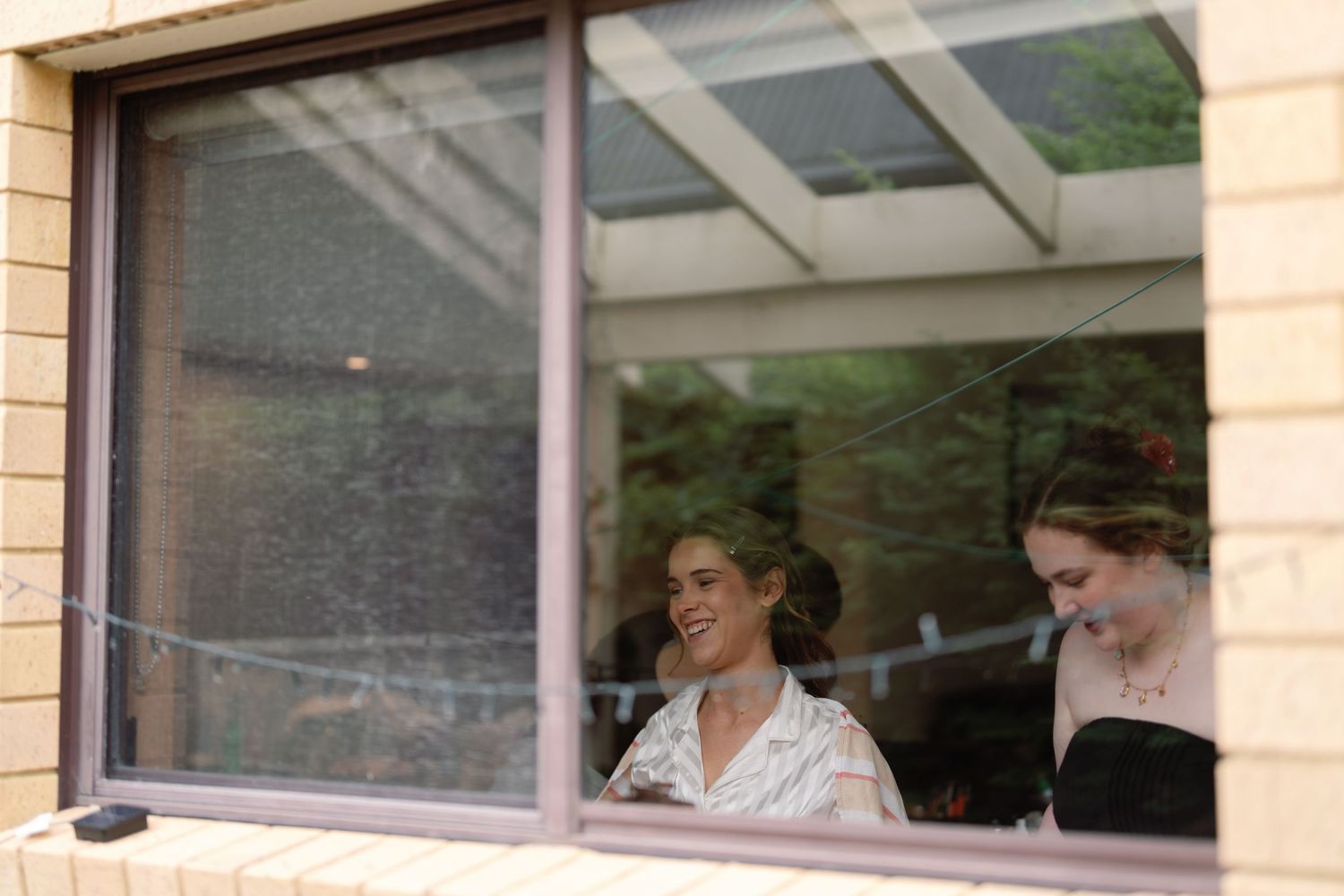 Two people are seen through a window frame, one wearing white and one in black, sharing a candid moment together.